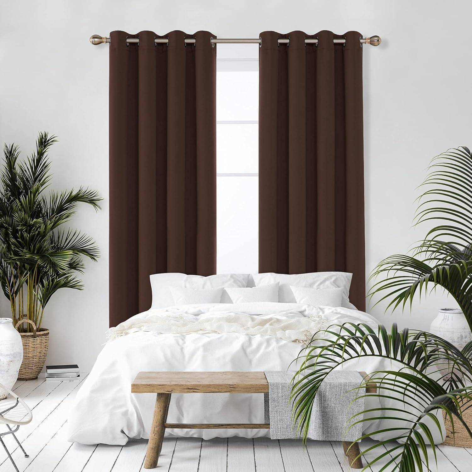 Ein gemütliches Schlafzimmer mit einer weißen Bettdecke, einer Holzbank am Fußende, braunen Vorhängen, die ein großes Fenster verdecken, und üppigen grünen Pflanzen rundherum.