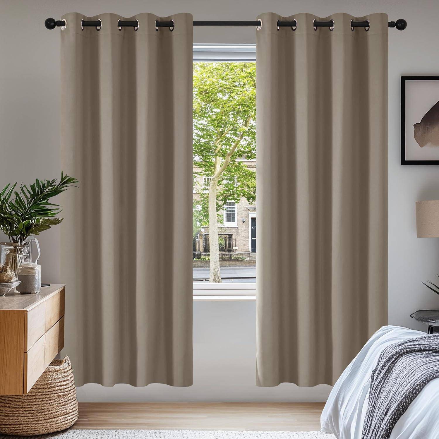 Ein Schlafzimmer mit beigen Vorhängen, einem Holznachttisch mit Büchern und einer Pflanze und einem Fenster, das einen Blick auf eine baumgesäumte Straße freigibt.