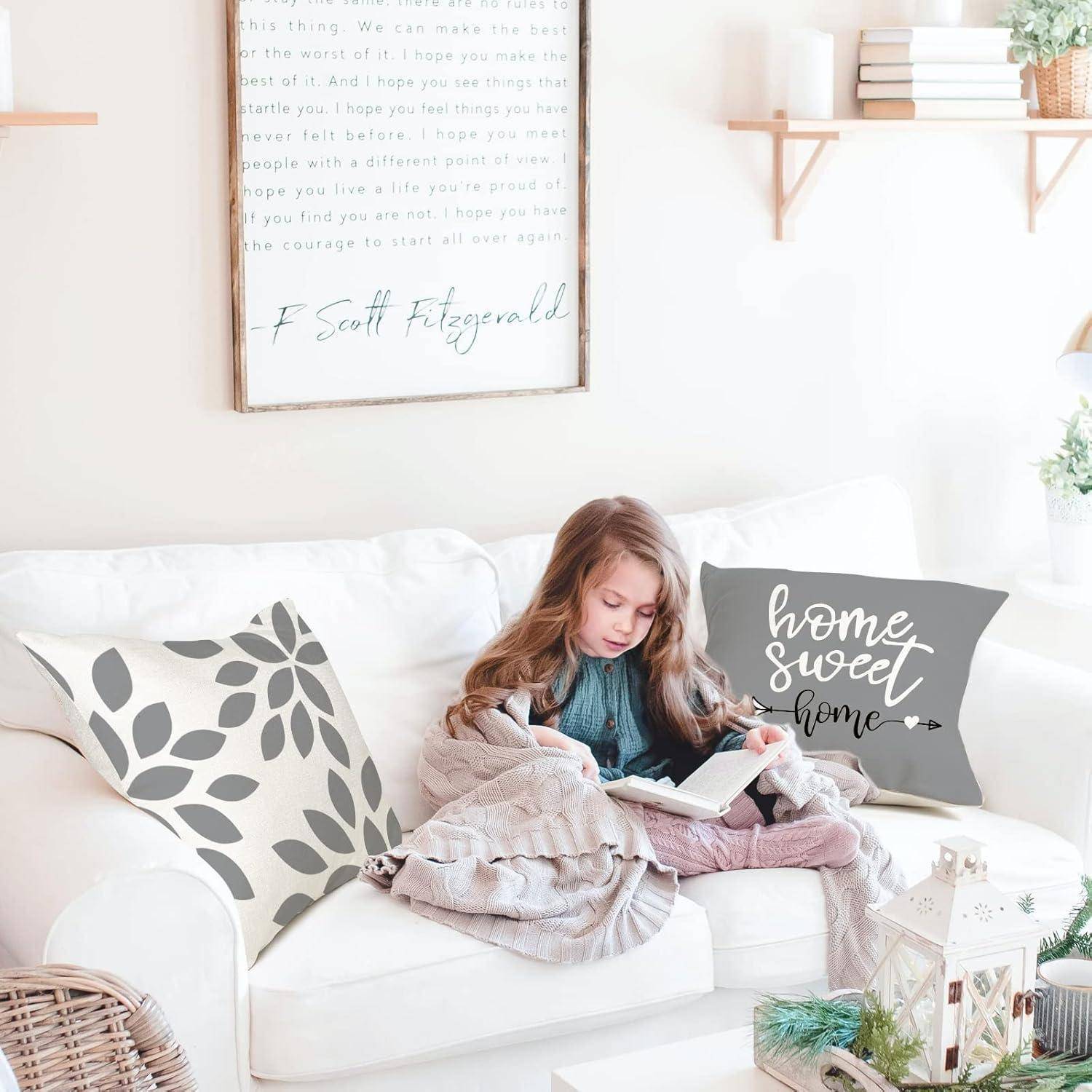 Ein junges Mädchen sitzt auf einem weißen Sofa und liest ein Buch. Kissen, eines mit Blattmustern und ein anderes mit dem Schriftzug ‚home sweet home', umgeben sie.