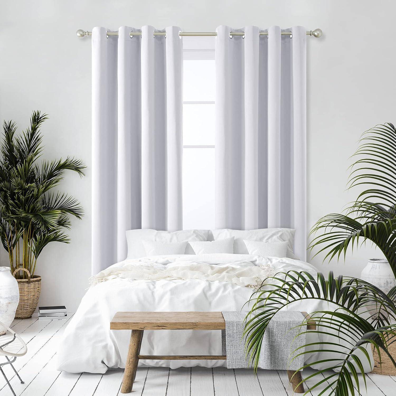 Ein gemütliches Schlafzimmer präsentiert ein ordentlich gemachtes Bett mit weißen Bettwäsche, flankiert von hohen grünen Pflanzen. Sonnenlicht fällt durch weiße Gardinen.