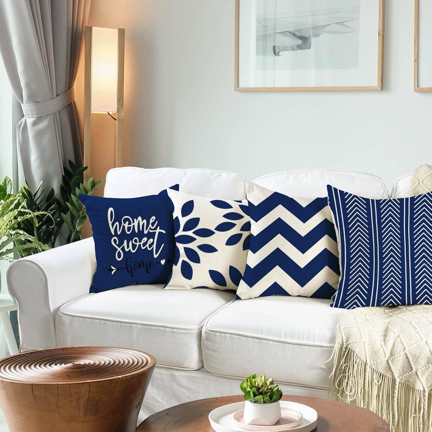Ein weißes Sofa mit vier dekorativen Kissen in Navy-Blau und weißen Mustern; eines davon trägt die Aufschrift ‚home sweet home'. Eine Pflanze, eine Lampe und ein gerahmtes Kunstwerk in der Nähe.