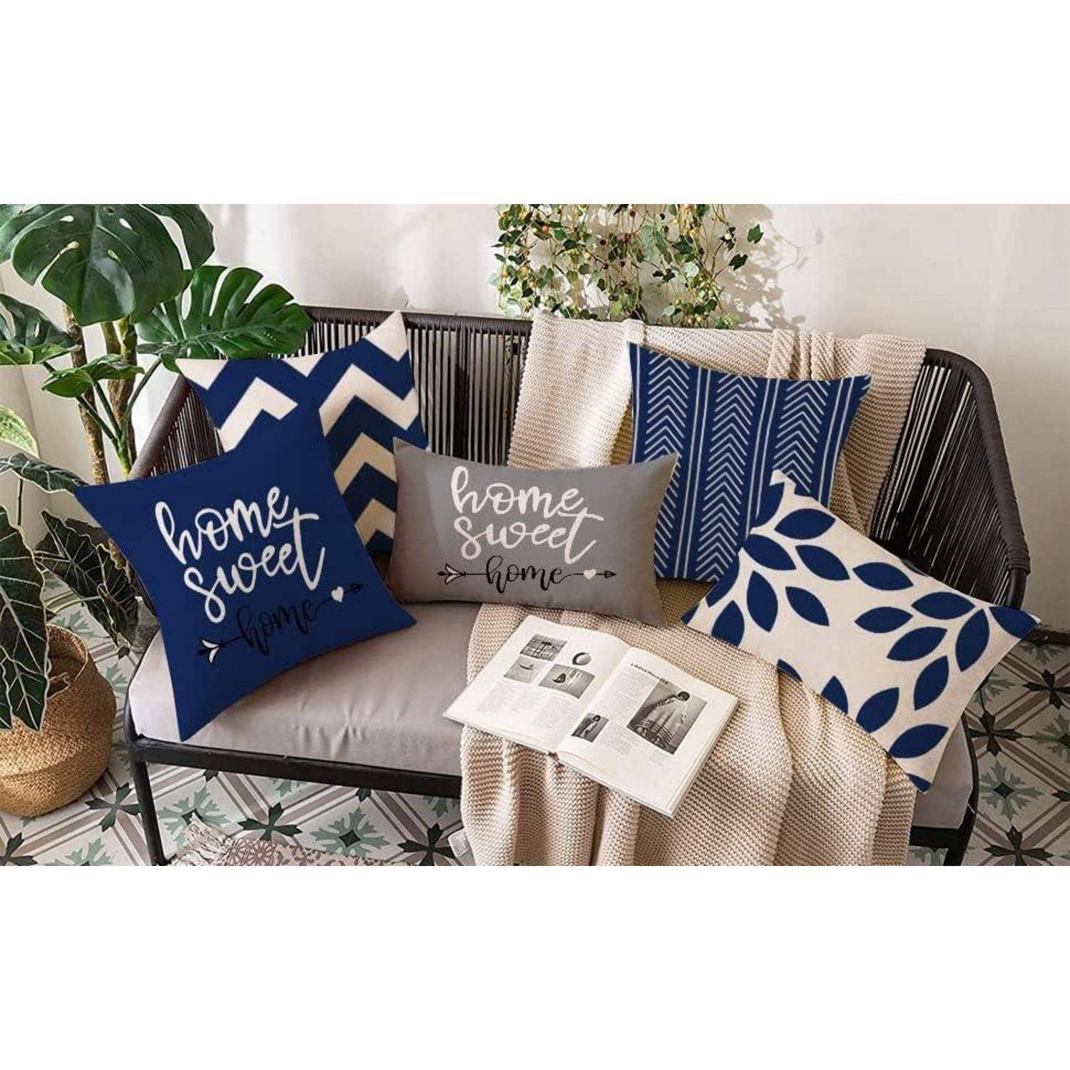 Eine gemütliche Couch mit einer Mischung aus blauen und grauen Kissen, auf denen der Text 'Home Sweet Home' zu sehen ist, umgeben von Pflanzen und einem aufgeschlagenen Buch auf einer Decke.