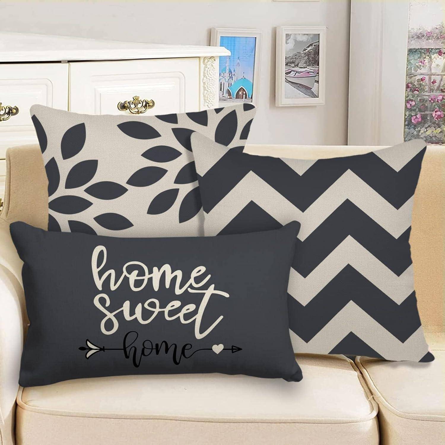 Drei Zierkissen auf einem beigen Sofa; eines mit dem Text ‚Zu Hause süß Zuhause', ein anderes mit einem Blattmuster und das dritte mit einem Zickzackmuster.