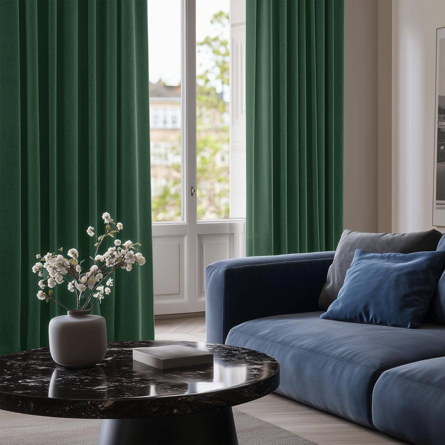 Ein moderner Wohnraum mit einem dunkel marmorierten Couchtisch, auf dem eine Vase mit weißen Blumen steht, einem blauen Sofa mit Kissen und großen grünen Vorhängen.