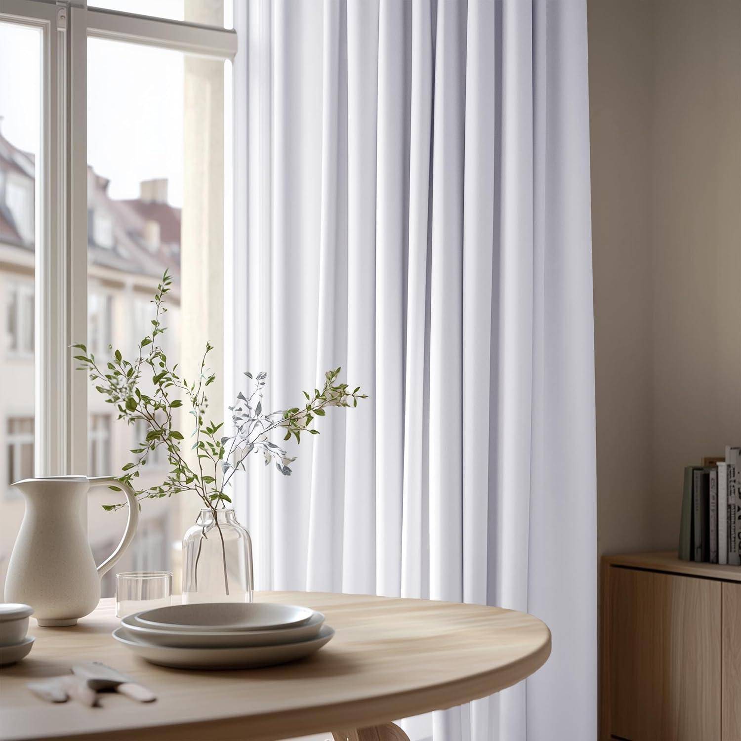 Ein runder Tisch neben einem Fenster trägt einen weißen Krug, eine Vase mit Zweigen und Teller, mit weißen Vorhängen und Blick auf ein Gebäude draußen.