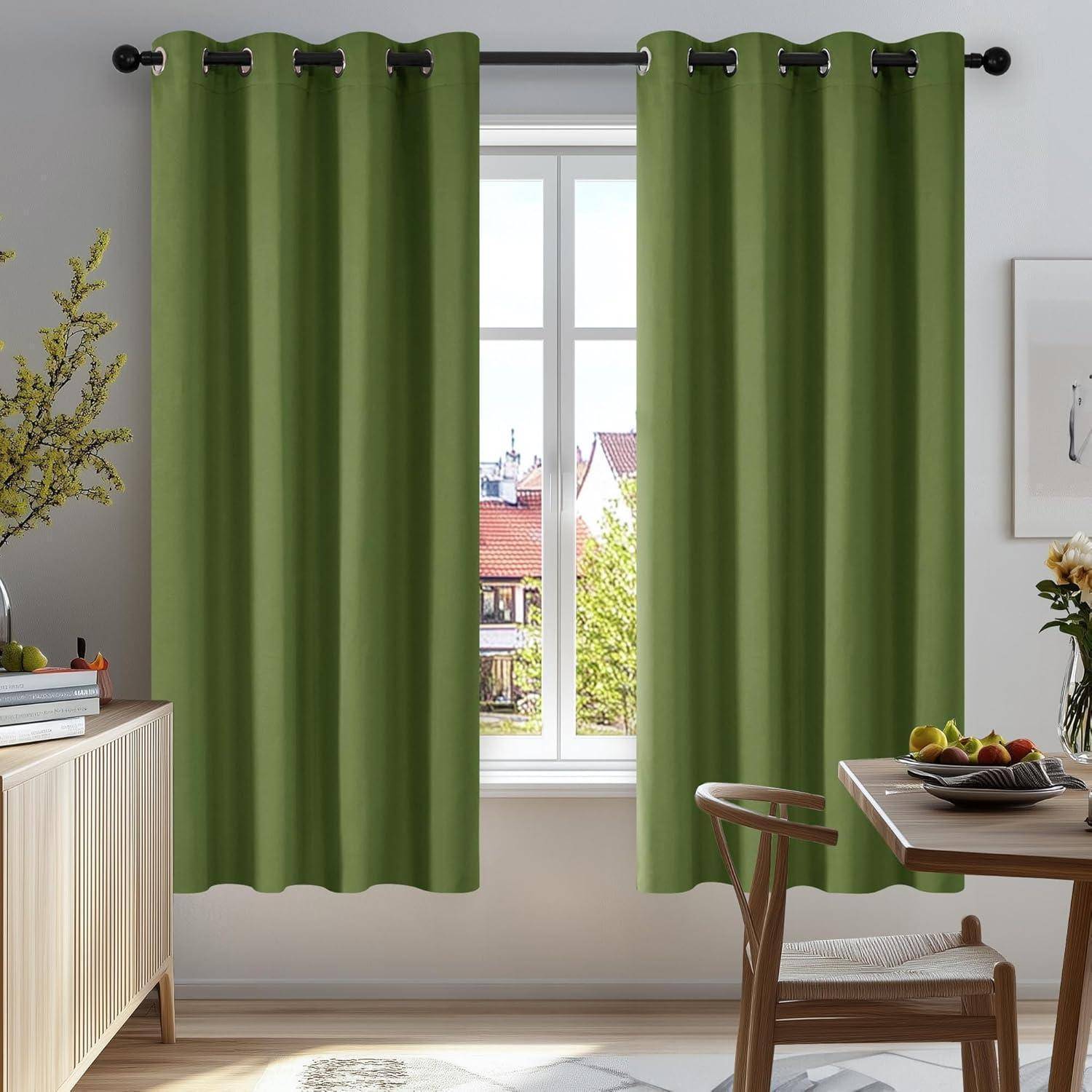 Ein von Sonnenlicht durchfluteter Raum mit grünen Vorhängen, ein Fensterblick auf Dächer und Bäume, ein Holzsideboard und ein Esstisch mit Obst und Pflanzen.