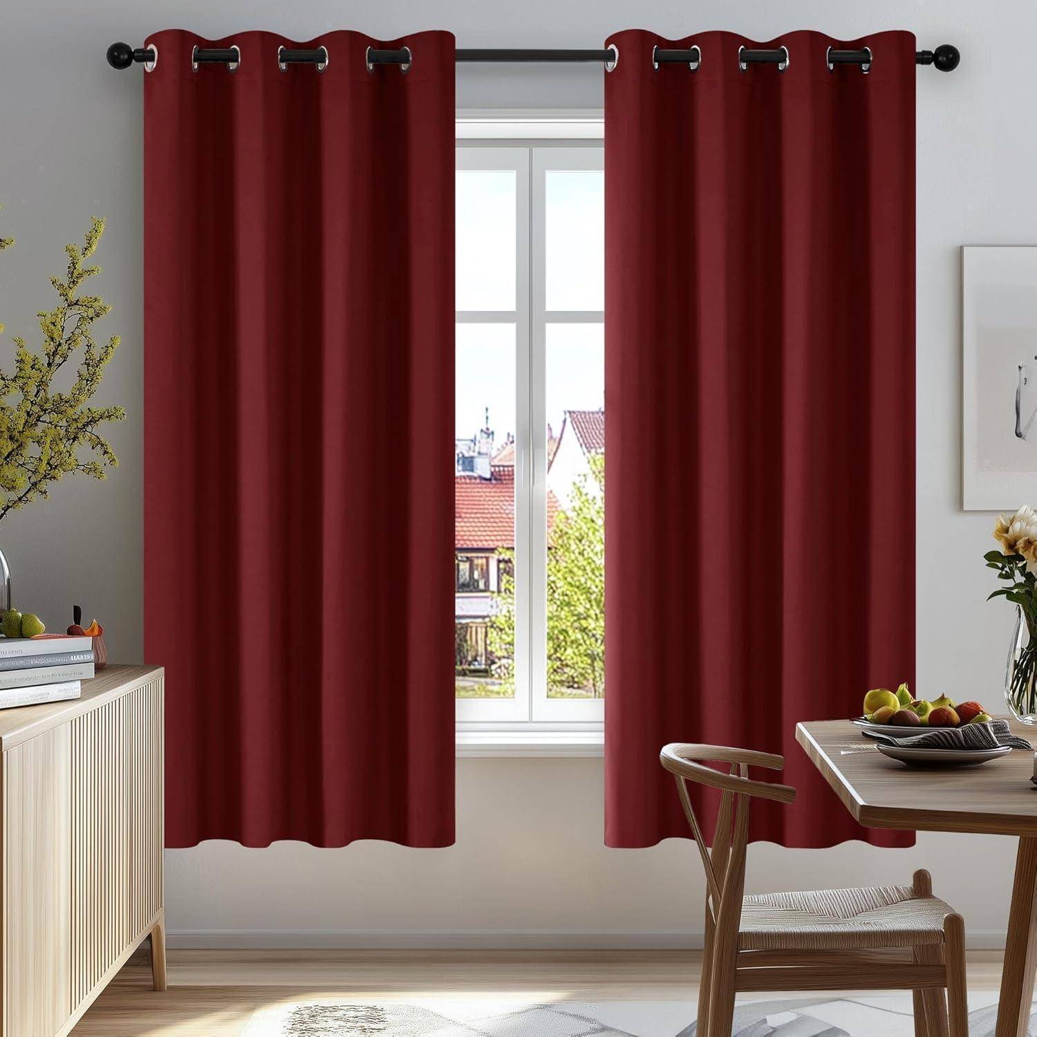Rote Vorhänge an einem Fenster mit Stadtblick, neben einem Tisch mit Büchern und einer Pflanze in einem sonnendurchfluteten Raum.