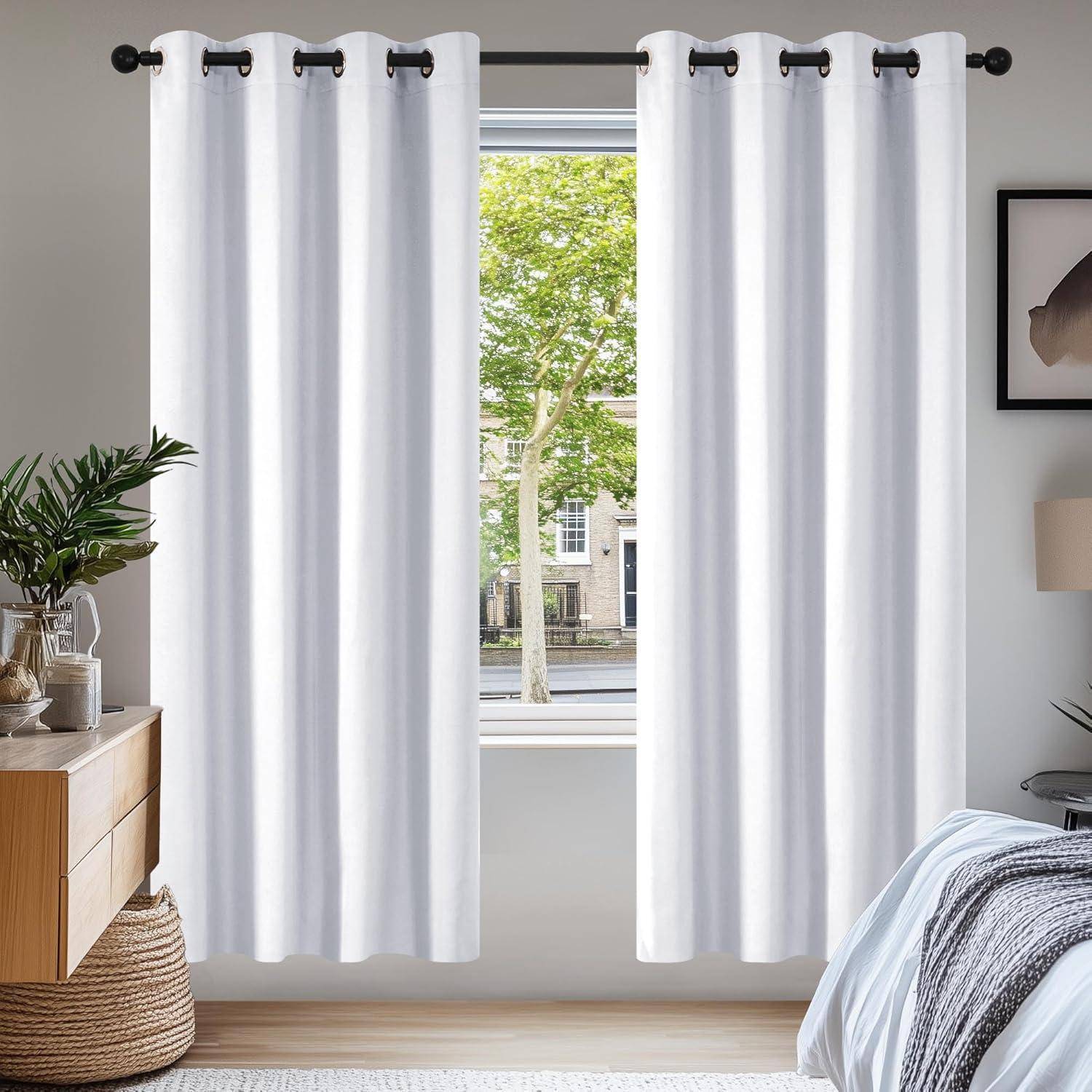 Weiße Vorhänge hängen an einer Stange vor einem Fenster, das eine baumgesäumte Straße zeigt. Das Interieur umfasst einen Holzschrank mit Dekoration.