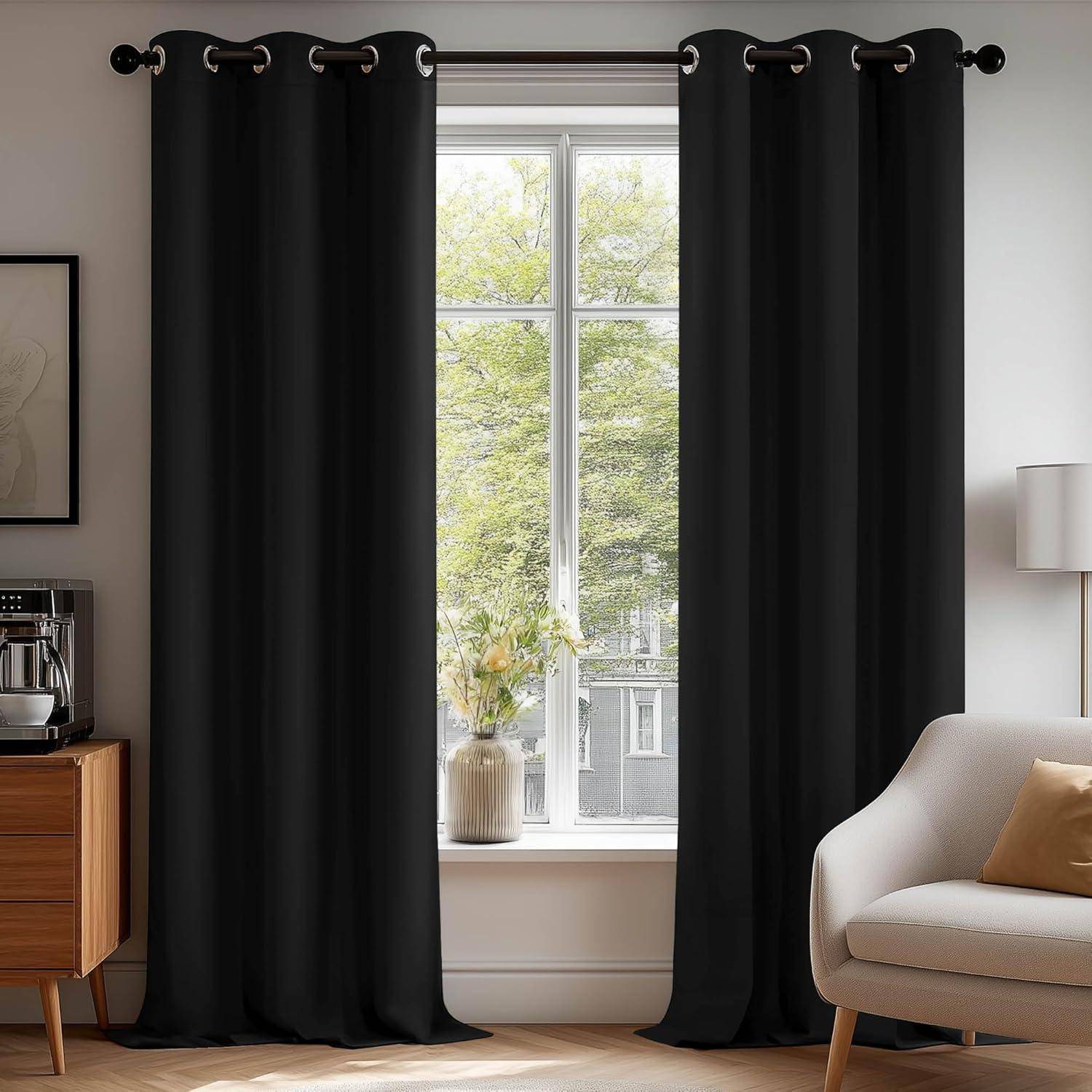 Ein heller Raum mit einem Fenster, das von schwarzen Vorhängen umrahmt ist und einen Blick auf grüne Bäume nach draußen freigibt. Ein hellbeiges Sofa und Holzmöbel ergänzen den Raum.