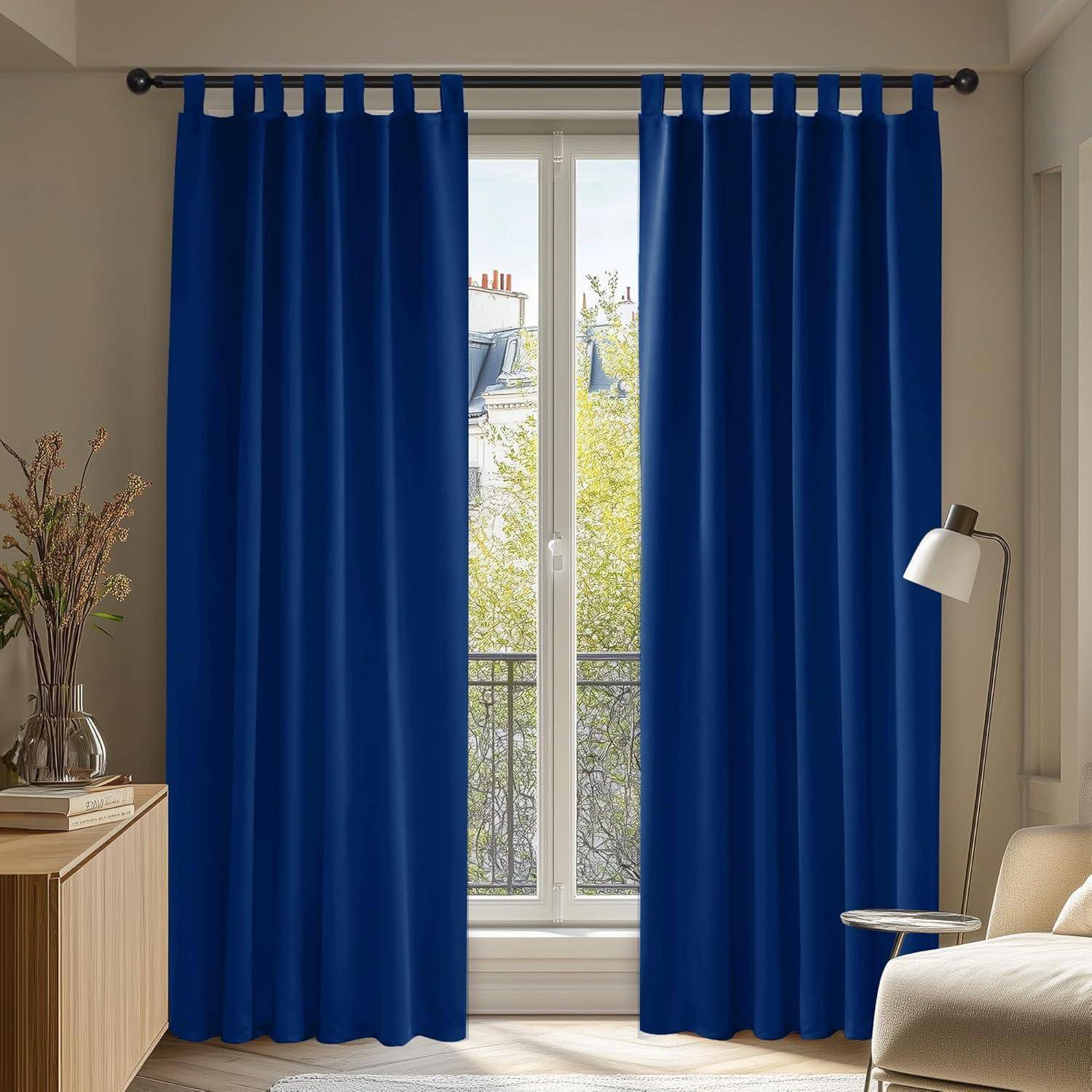 Ein gemütliches Zimmer zeigt blaue Vorhänge, die ein großes Fenster umrahmen, das einen Blick auf Bäume und Dächer freigibt. Eine Stehlampe steht neben einem Holzschrank.
