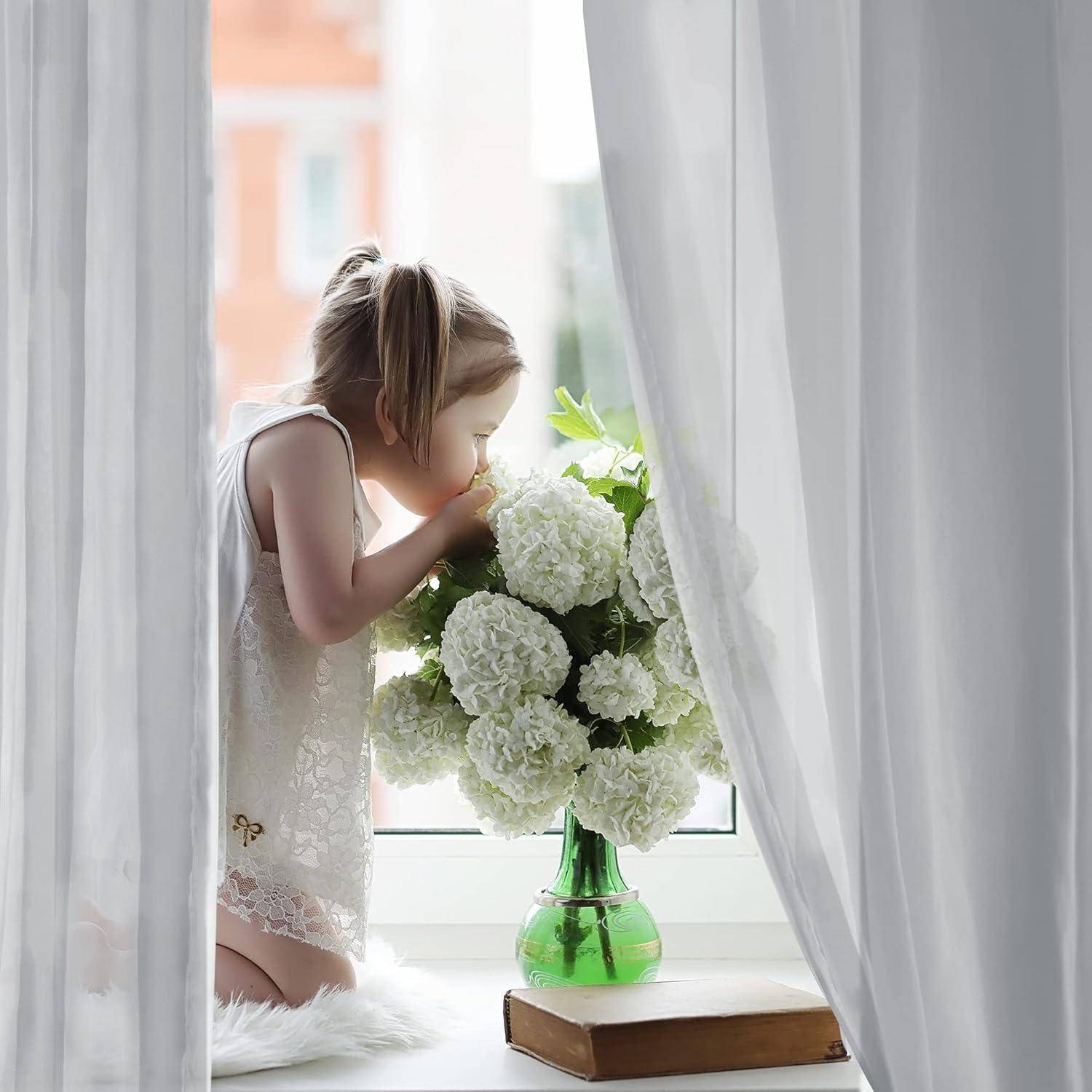 Ein junges Mädchen in einem weißen Kleid kniet an einem Fenster und riecht an einem Blumenstrauß in einer grünen Vase, während Sonnenlicht durch durchsichtige Gardinen fällt.