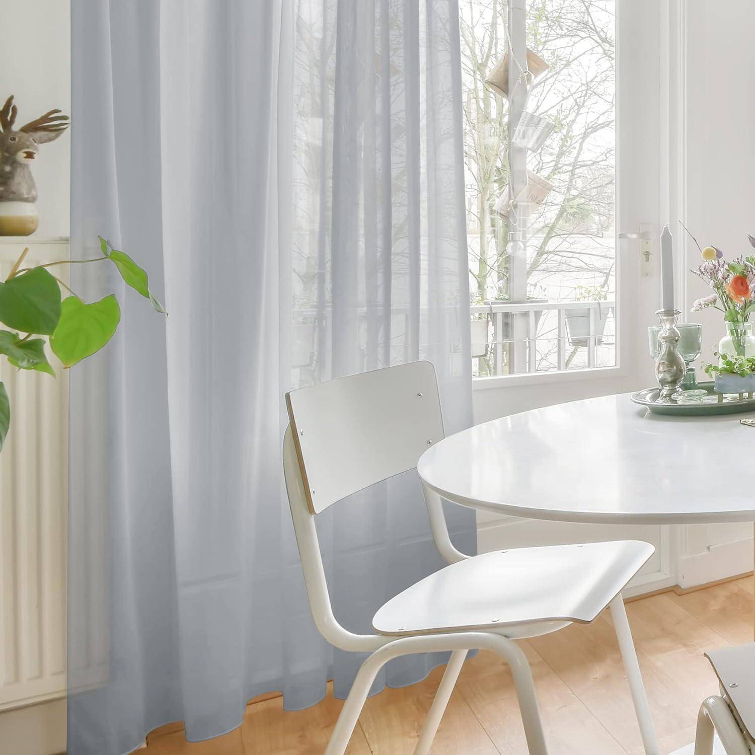 Ein sonnendurchfluteter Essbereich mit einem weißen Stuhl und einem runden Tisch in der Nähe eines Fensters mit transparenten Vorhängen, der Blick geht auf eine baumgesäumte Straße.
