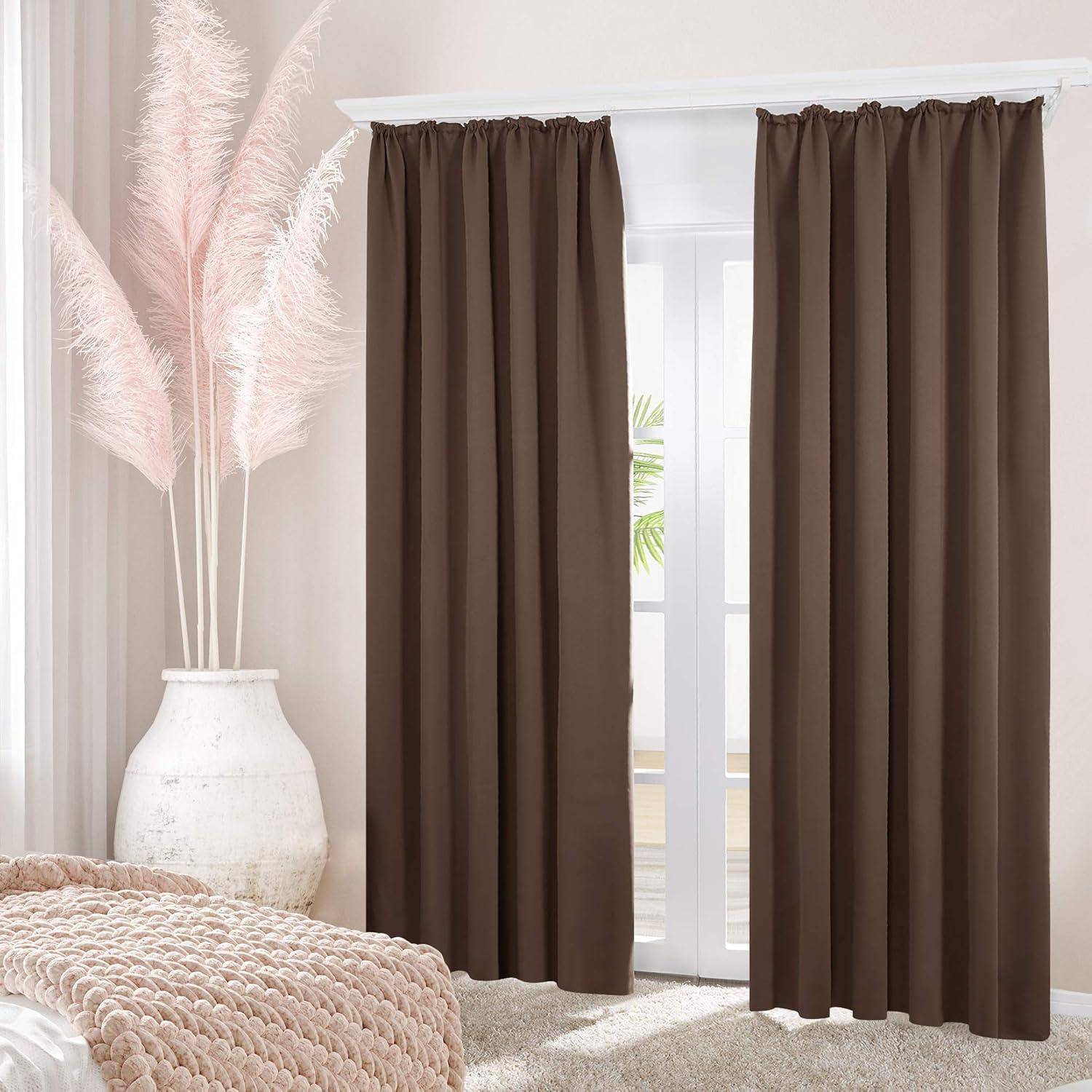 Ein Raum zeigt ein großes Fenster mit braunen Vorhängen und eine weiße Keramikvase, die hohe, federartige rosa Dekorationen neben einem beigen Bett enthält.
