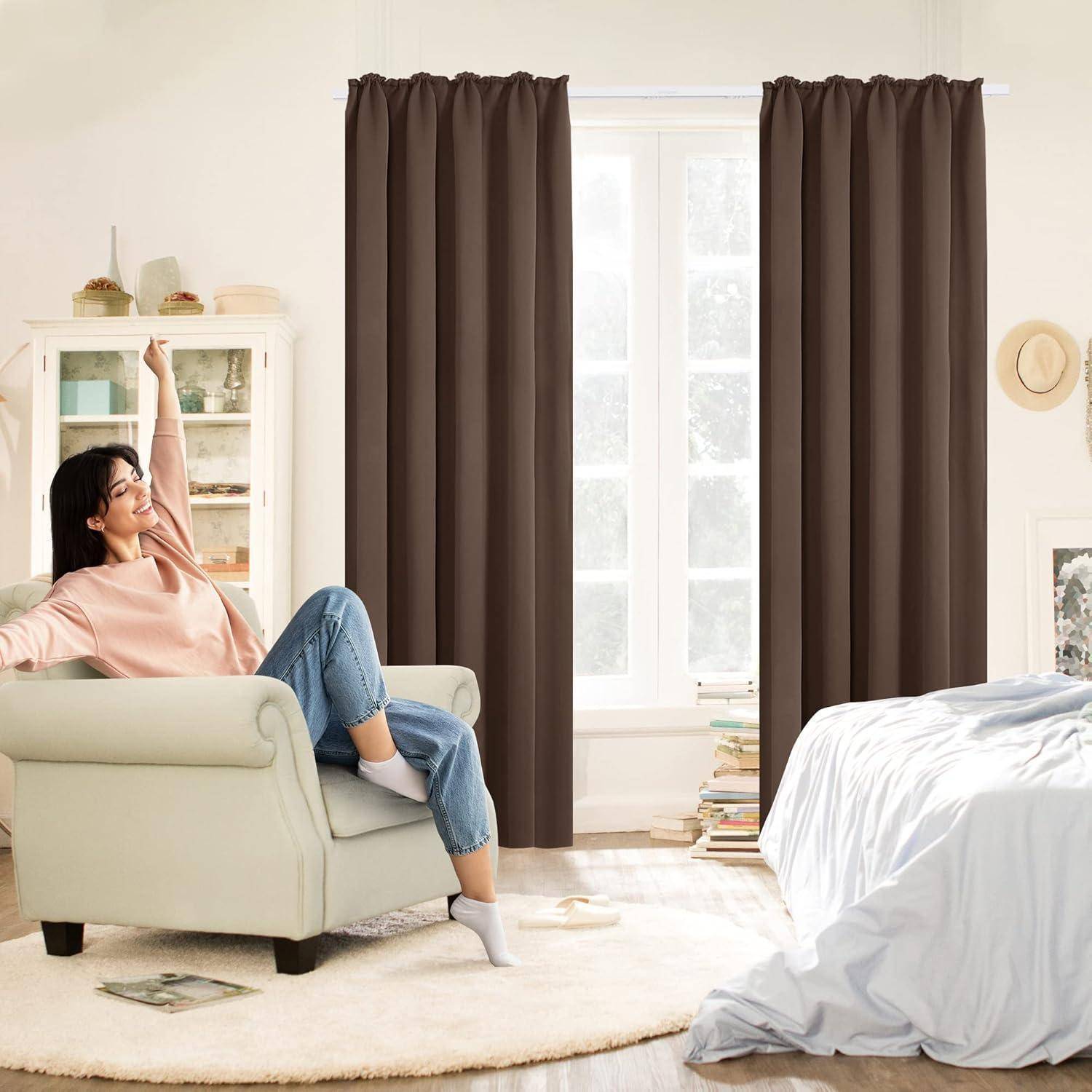 Ein gemütliches Schlafzimmer mit hellen Wänden und einem Bett mit weißer Bettwäsche. Eine Person lächelt, während sie sich in einem Sessel streckt. Braune Vorhänge bedecken zwei hohe Fenster.