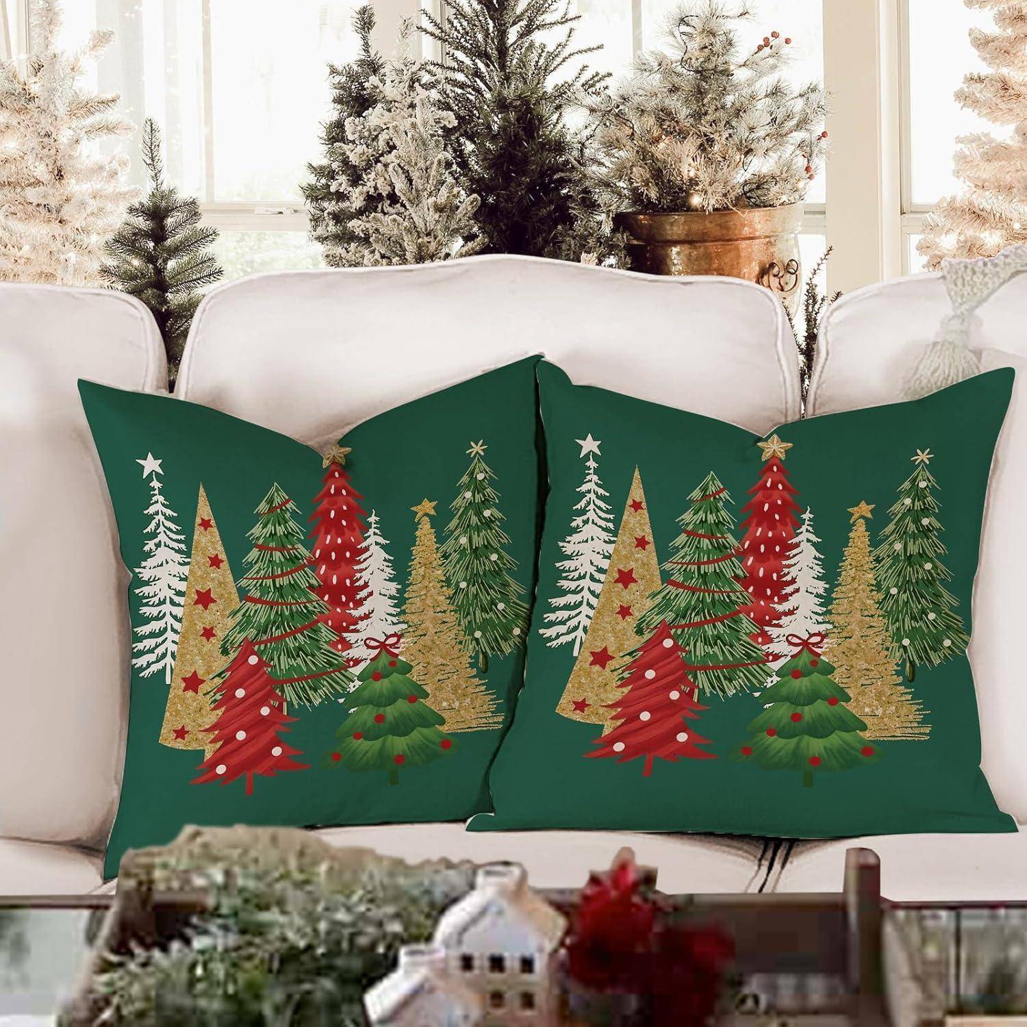 Weihnachtlich gestaltete Kissen mit verschiedenen geschmückten Weihnachtsbäumen liegen auf einem weißen Sofa, umgeben von verschneiten Zierbäumen.