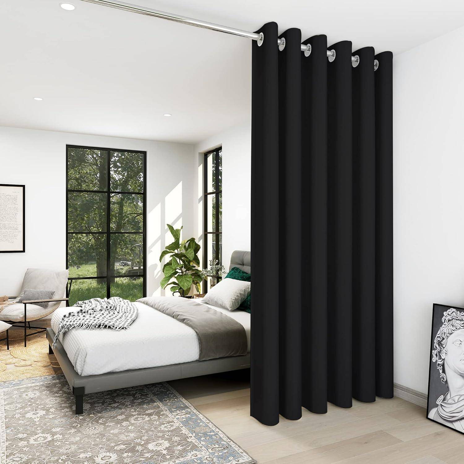 Schlafzimmer mit großen Fenstern, weißen Wänden und einem Bett mit grauer Bettwäsche. Es gibt einen Teppich, eine Pflanze und dunkle, bodenlange Vorhänge.
