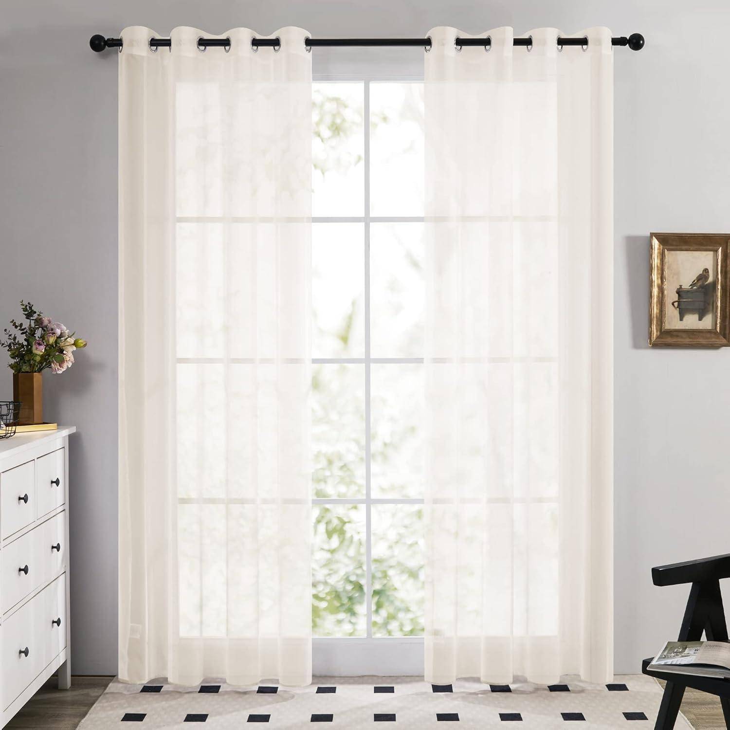 Heller Raum mit durchsichtigen weißen Gardinen an einem großen Fenster, einem schwarz-weißen Teppich, einer weißen Kommode mit Blumen und Wandkunst.