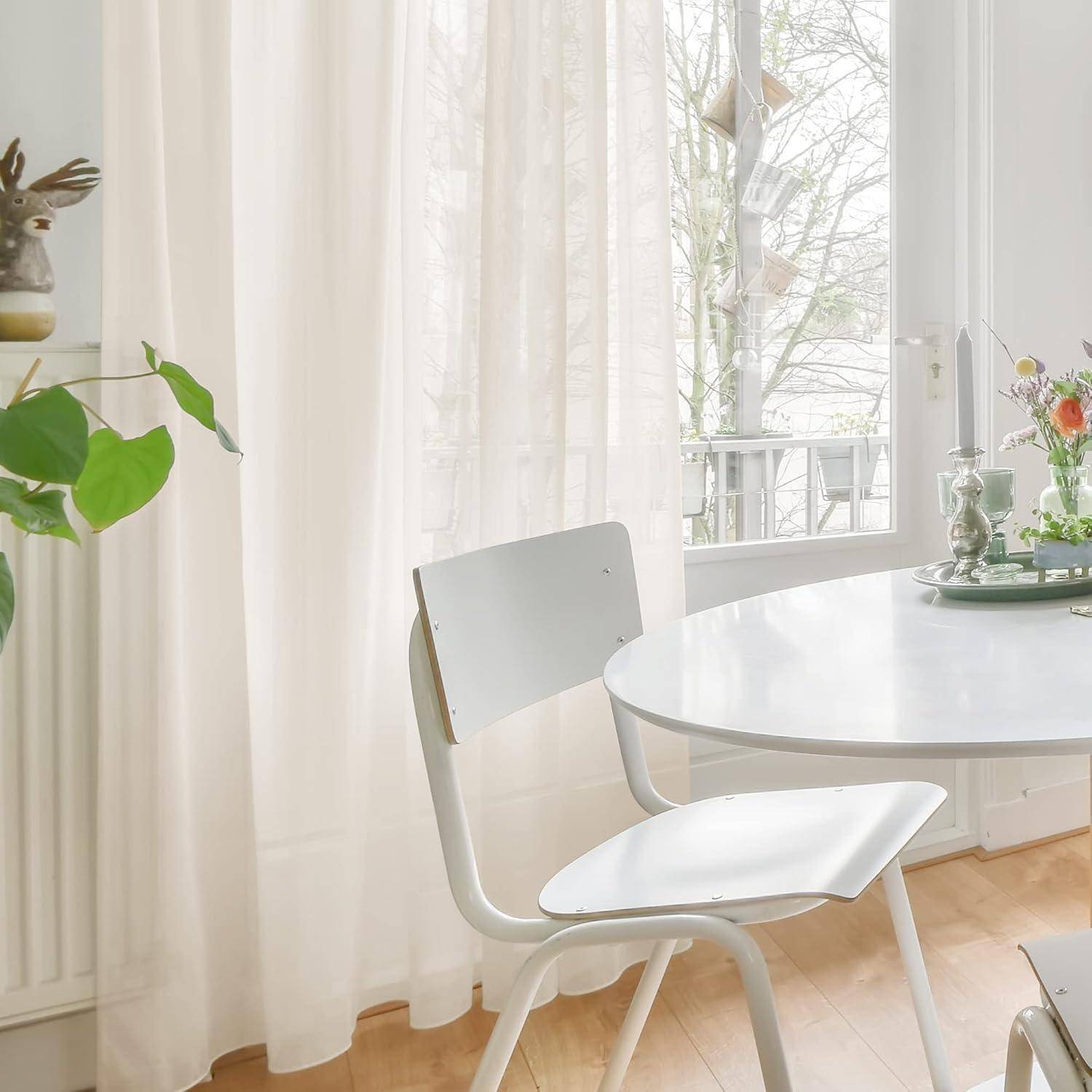 Ein heller, minimalistischer Essbereich mit einem weißen Tisch und einem Stuhl neben einem Fenster, geschmückt mit einer Topfpflanze und einem Kerzenhalter, blickt auf einen Balkon.