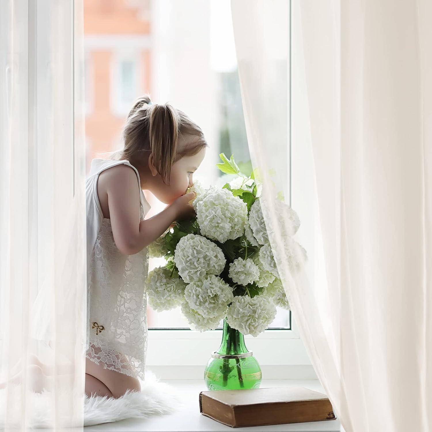 Ein junges Mädchen in einem weißen Kleid kniet auf der Fensterbank und riecht an einer Vase mit weißen Blumen, während weiches, natürliches Licht die Szene beleuchtet.