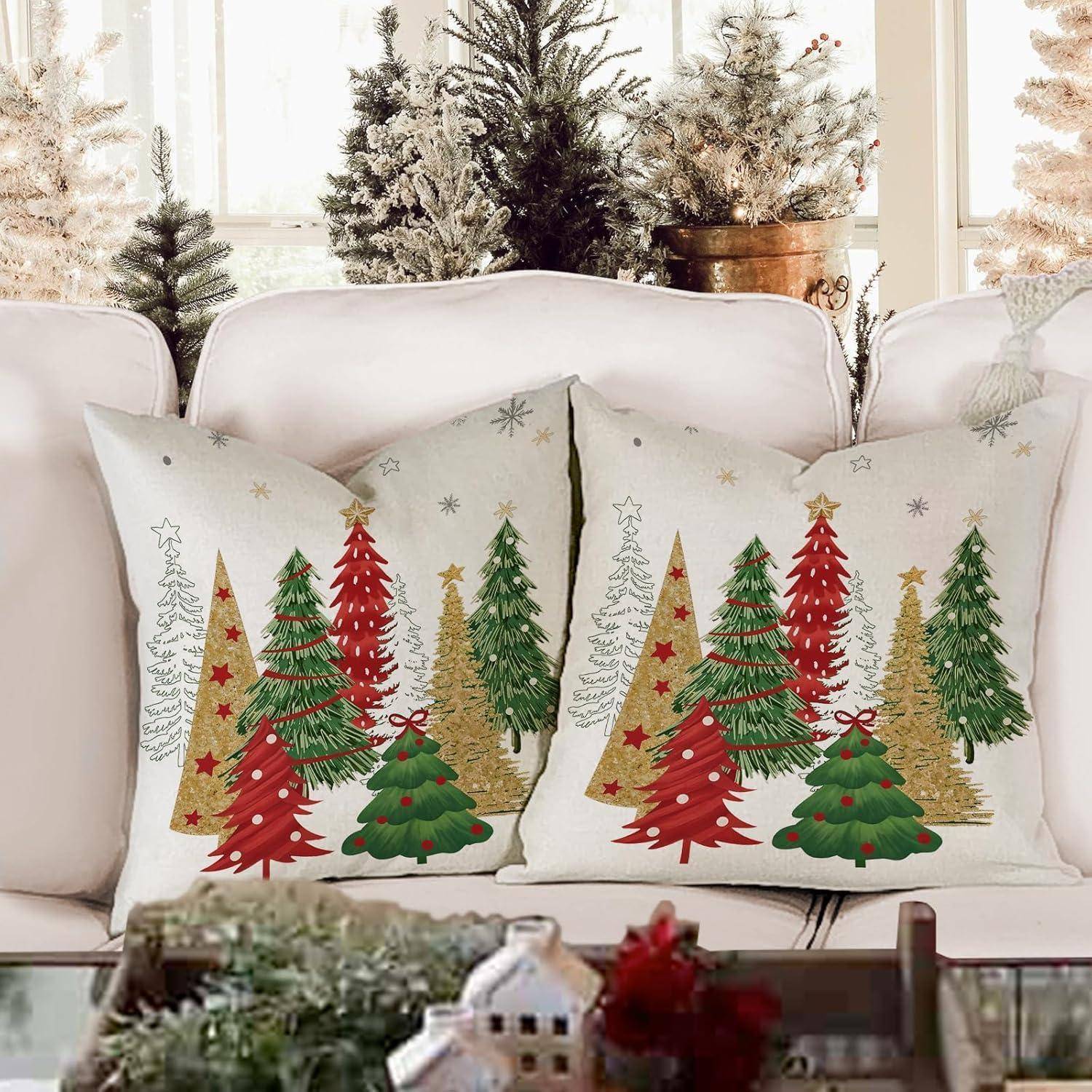 Weihnachtlich dekorierte Kissen mit geschmückten Bäumen liegen auf einem weißen Sofa; verschneite Topfbäume befinden sich im Hintergrund.