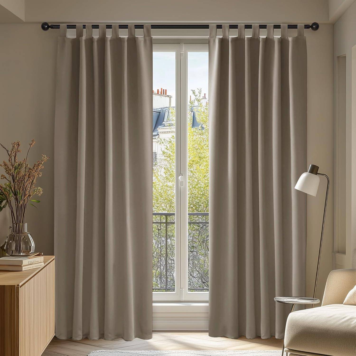 Ein sonniger Raum mit beigen Vorhängen, die über einem Fenster zugezogen sind und einen Blick auf Gebäude freigeben. Eine gemütliche Umgebung mit einer Lampe und einem Schrank in der Nähe.