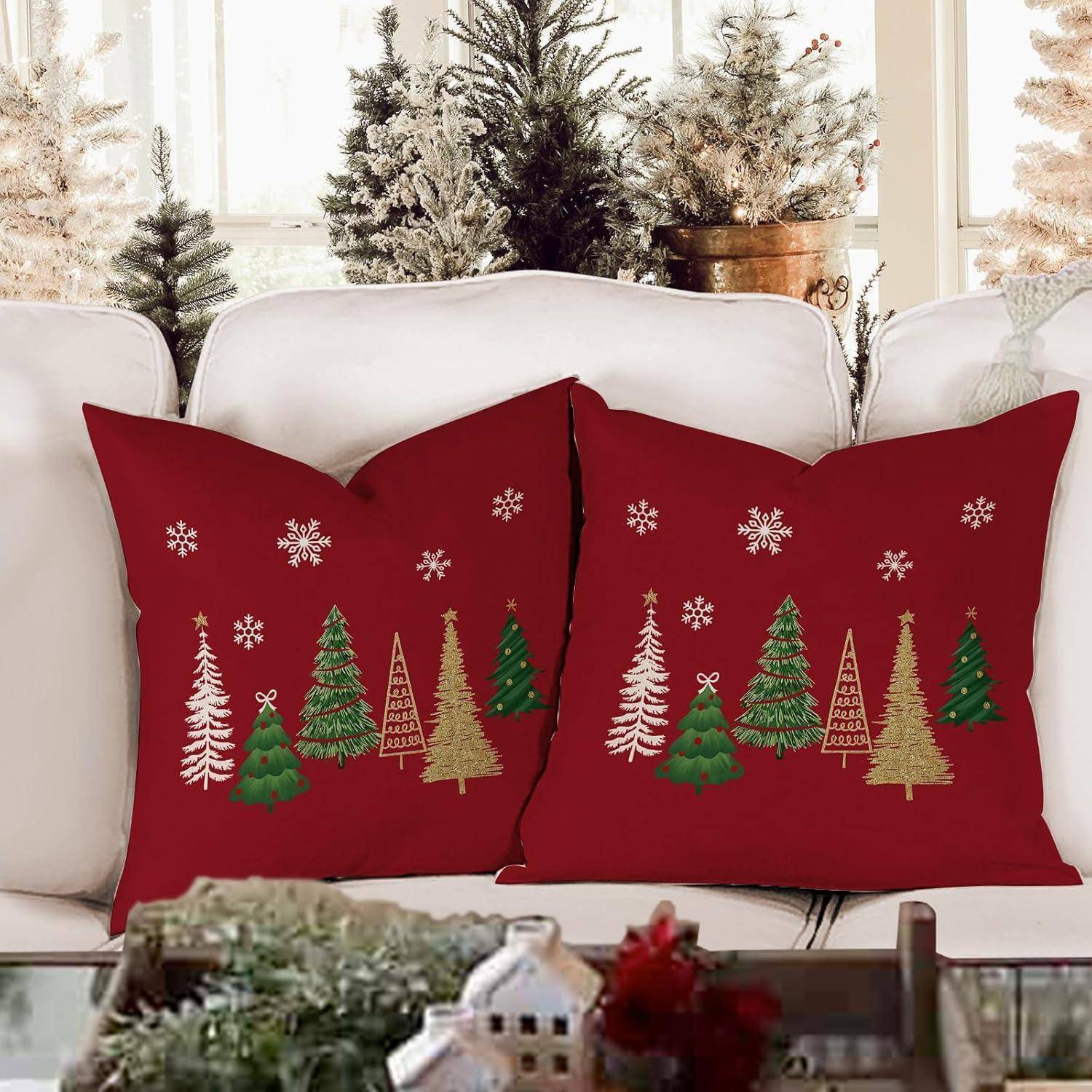 Rote Zierkissen mit festlichen Baum- und Schneeflockenmustern auf einem weißen Sofa mit verschneiten Bäumen im Hintergrund.