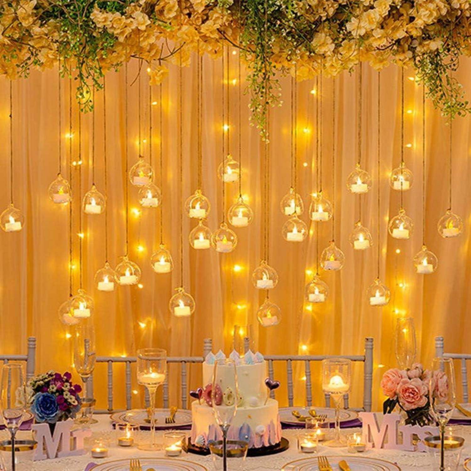 Hochzeitstisch mit Blumen- und Kerzendekorationen, auf dem eine Torte und die Aufschrift 'Mr & Mrs' platziert sind. Hängende Lichter erzeugen eine warme Atmosphäre.