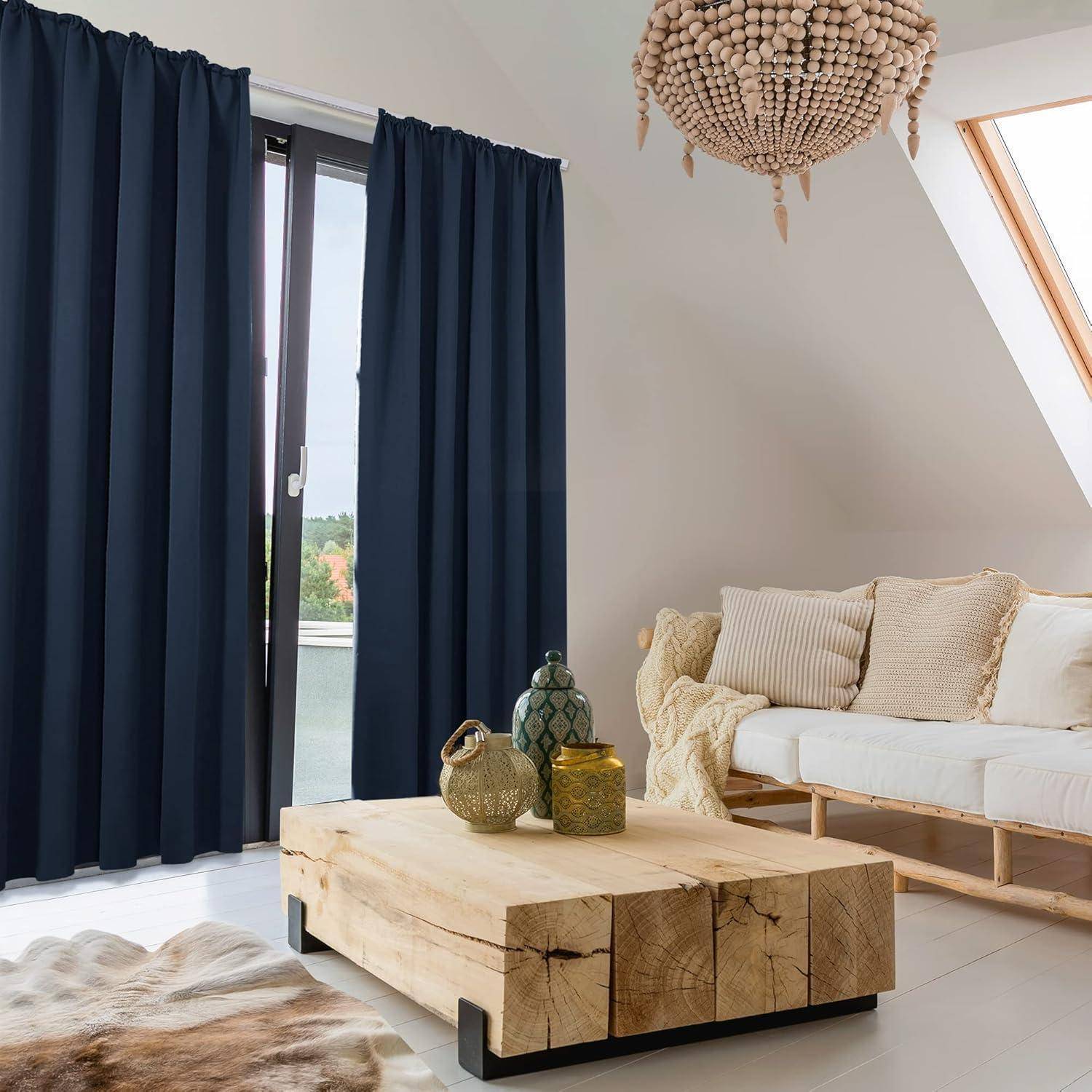 Ein moderner Wohnraum mit einem großen Fenster, das von dunklen Vorhängen verhangen ist, einem cremefarbenen Sofa mit Kissen und einem Holzkaffeetisch mit Vasen.
