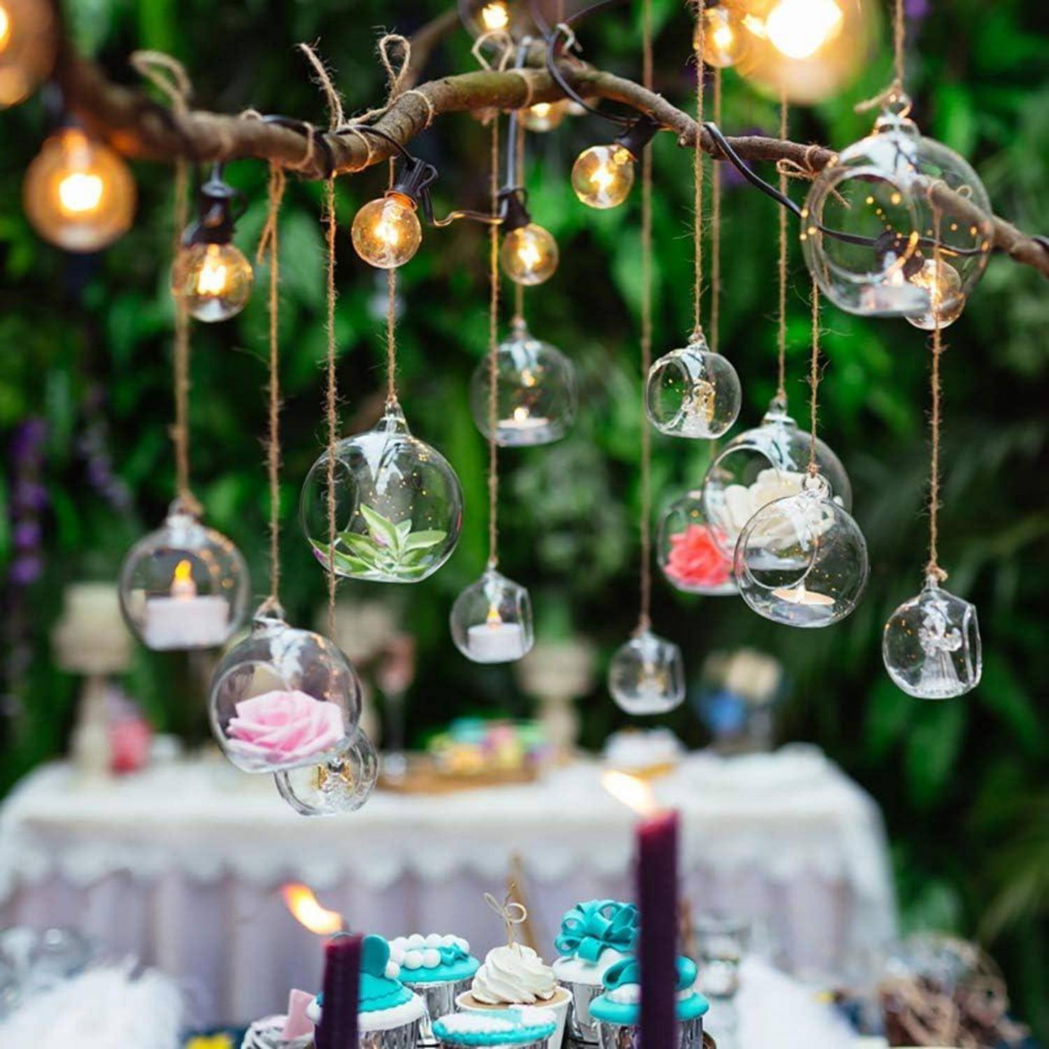 Hängende Glaskugeln mit Kerzen und Blumen schaffen eine zauberhafte Atmosphäre bei einer Outdoor-Party, wobei im Hintergrund ein dekorierter Tisch zu sehen ist.
