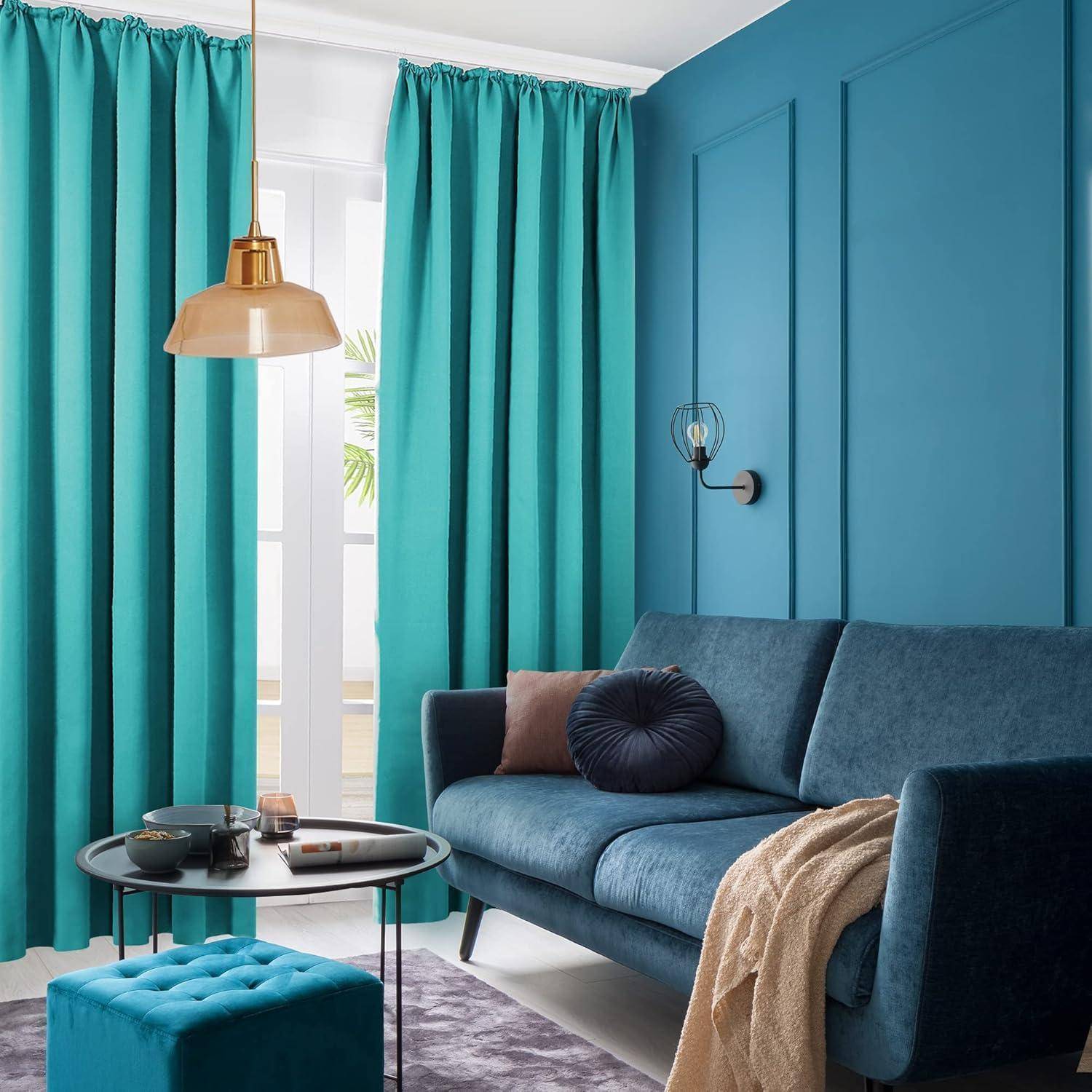 Ein modernes Wohnzimmer mit Vorhängen in Petrolblau, einer goldenen Pendelleuchte und einem blauen Sofa mit Kissen. Ein kleiner runder Tisch mit Büchern und einer Pflanze steht davor.