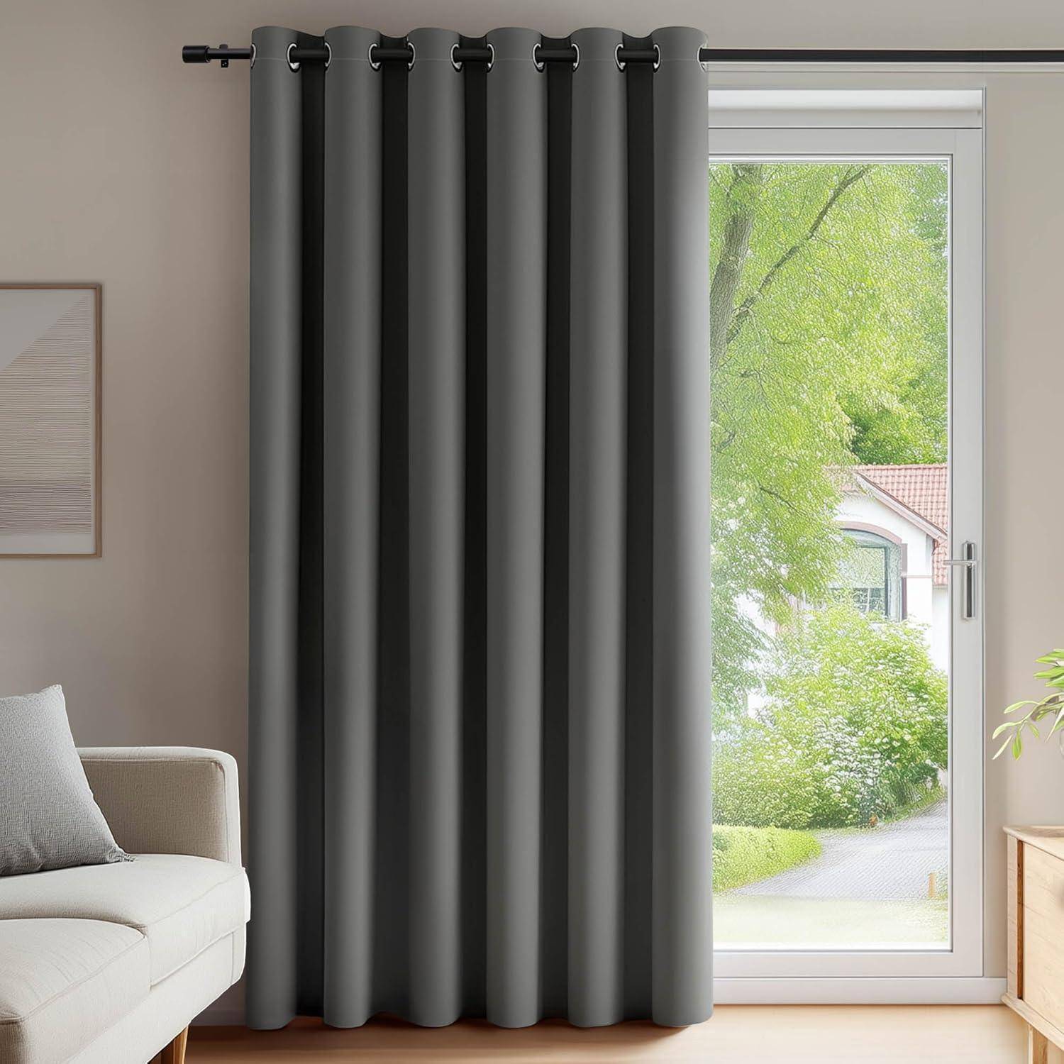 Ein dunkelgrauer Vorhang hängt von einer Stange und bedeckt ein hohes Fenster, das teilweise geöffnet ist und Blick auf Grünpflanzen und ein Haus draußen freigibt.