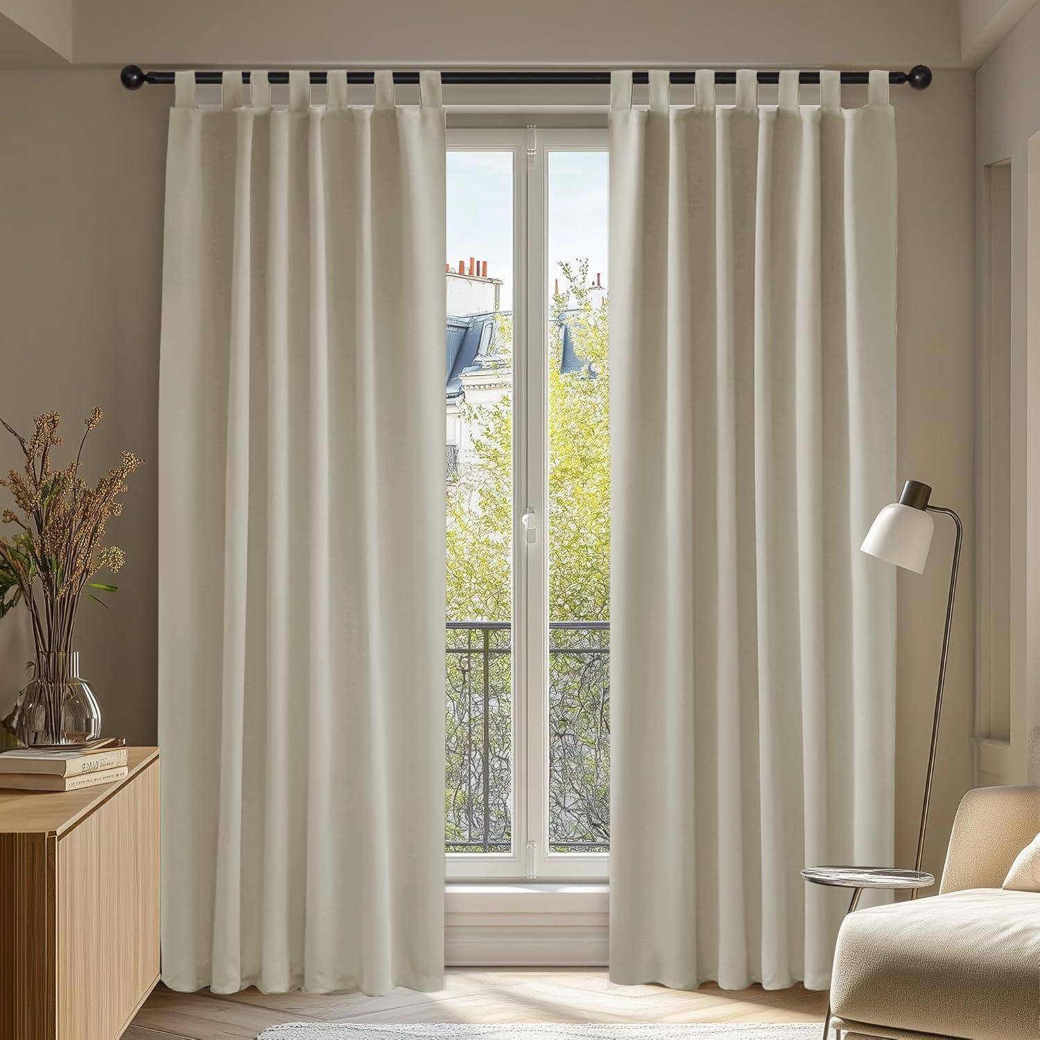 Ein sonnendurchfluteter Raum mit hellfarbenen bodenlangen Vorhängen, die ein großes Fenster umrahmen, mit Blick über Dächer. Ein kleiner Tisch und eine Lampe stehen in der Nähe.