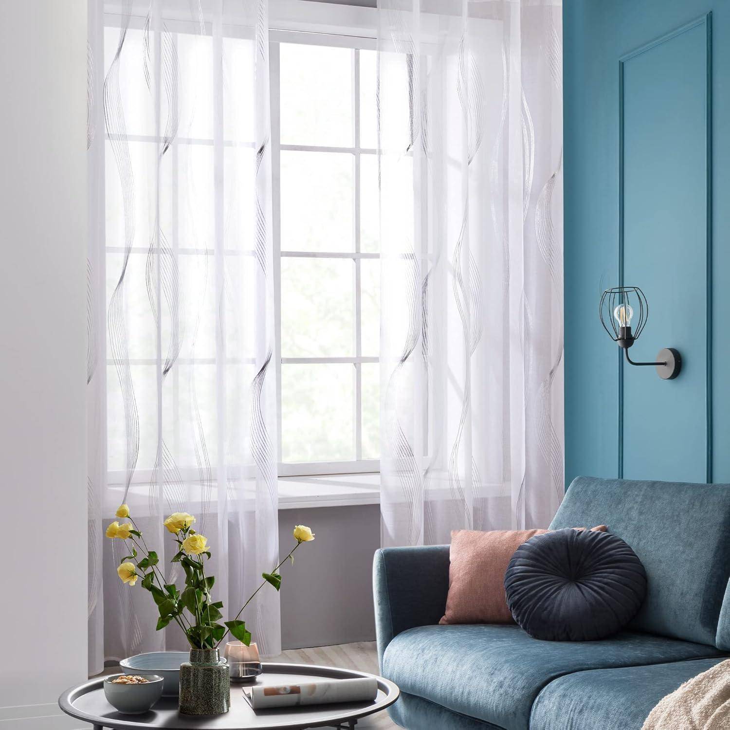 Ein gemütliches Wohnzimmer zeigt ein blaues Sofa mit einem runden Kissen, durchsichtige Gardinen, Topfpflanzen und einen kleinen Tisch neben einem Fenster.