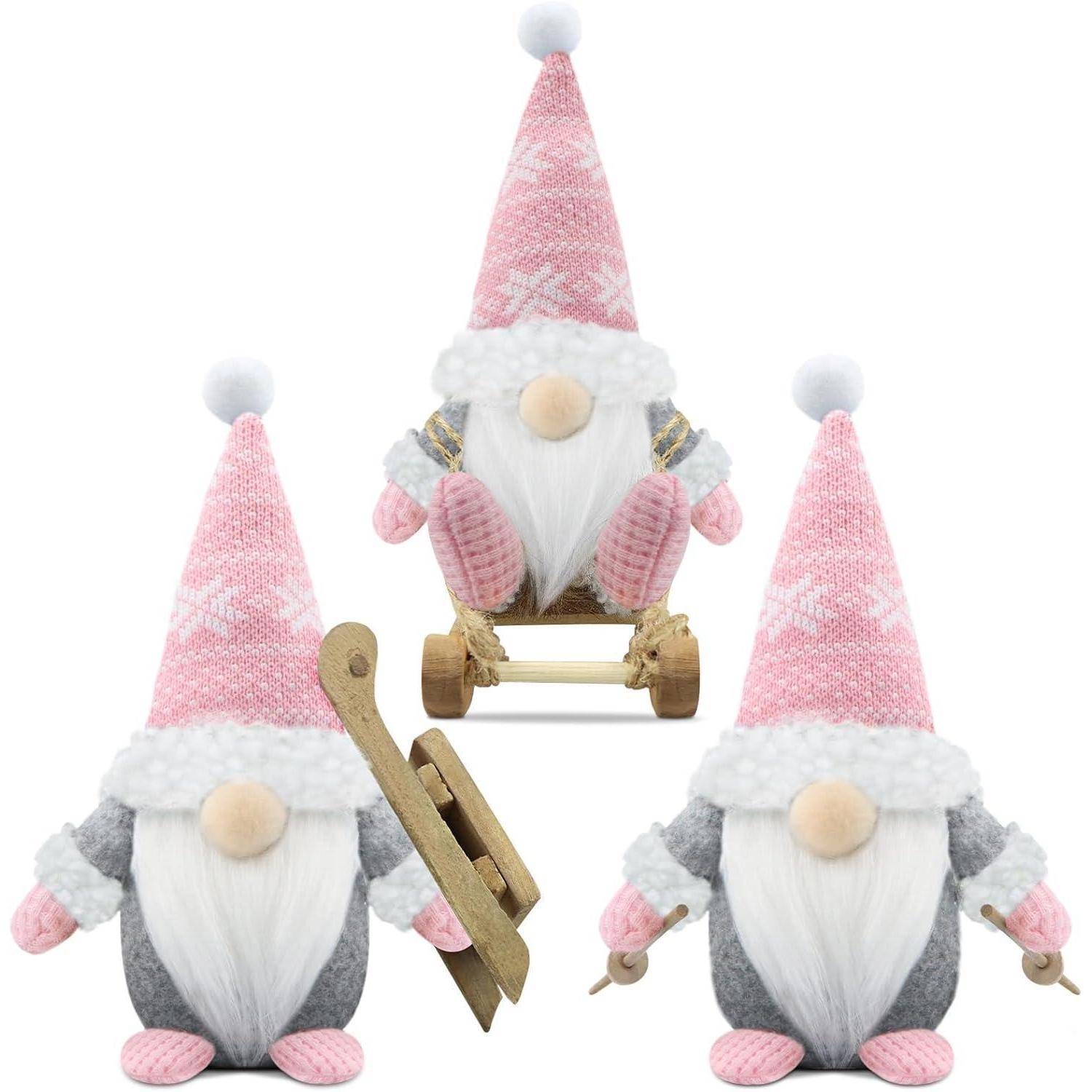 Drei Plüsch-Gnome in grauer Kleidung mit rosa Mützen. Zwei halten Holzschlitten, einer sitzt auf einem dritten Schlitten. Jeder trägt einen flauschigen weißen Besatz.