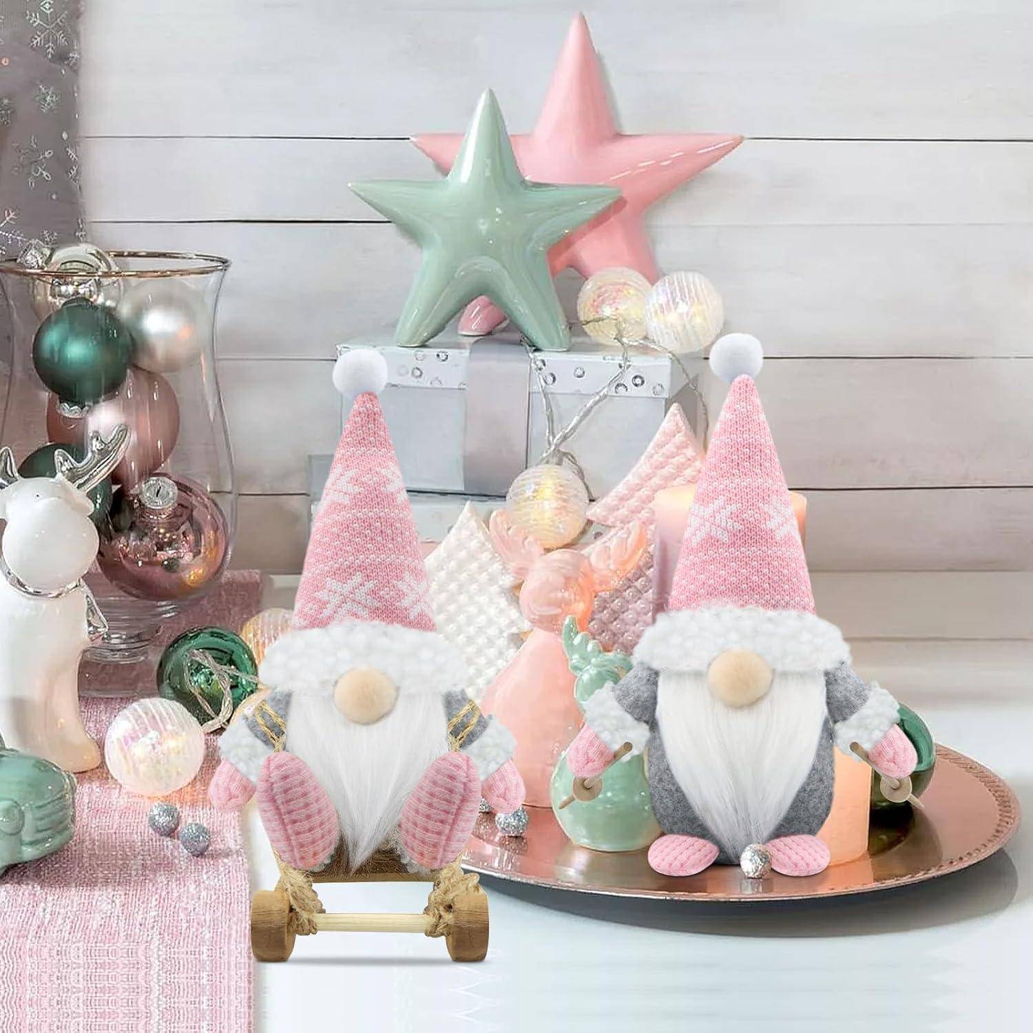 Zwei rosa und graue Feiertagszwerge sitzen auf einem Tisch, der mit pastellfarbenen Weihnachtsschmuck, darunter Sterne und Christbaumkugeln, in sanftem Licht dekoriert ist.