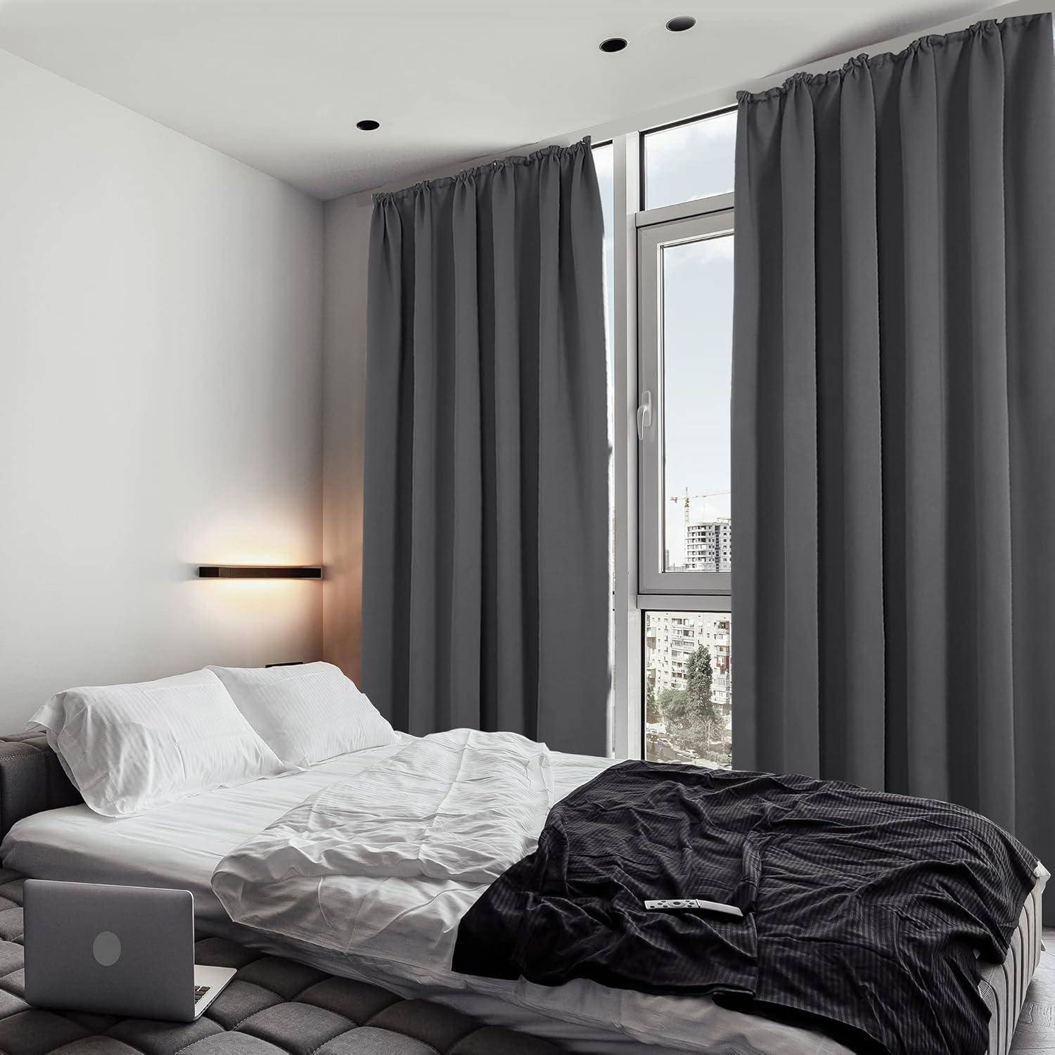 Ein modernes Schlafzimmer mit einem ordentlich gemachten Bett, grauen Vorhängen, die ein hohes Fenster verdecken, und weichem Ambiente-Licht von einer wandmontierten Leuchte.