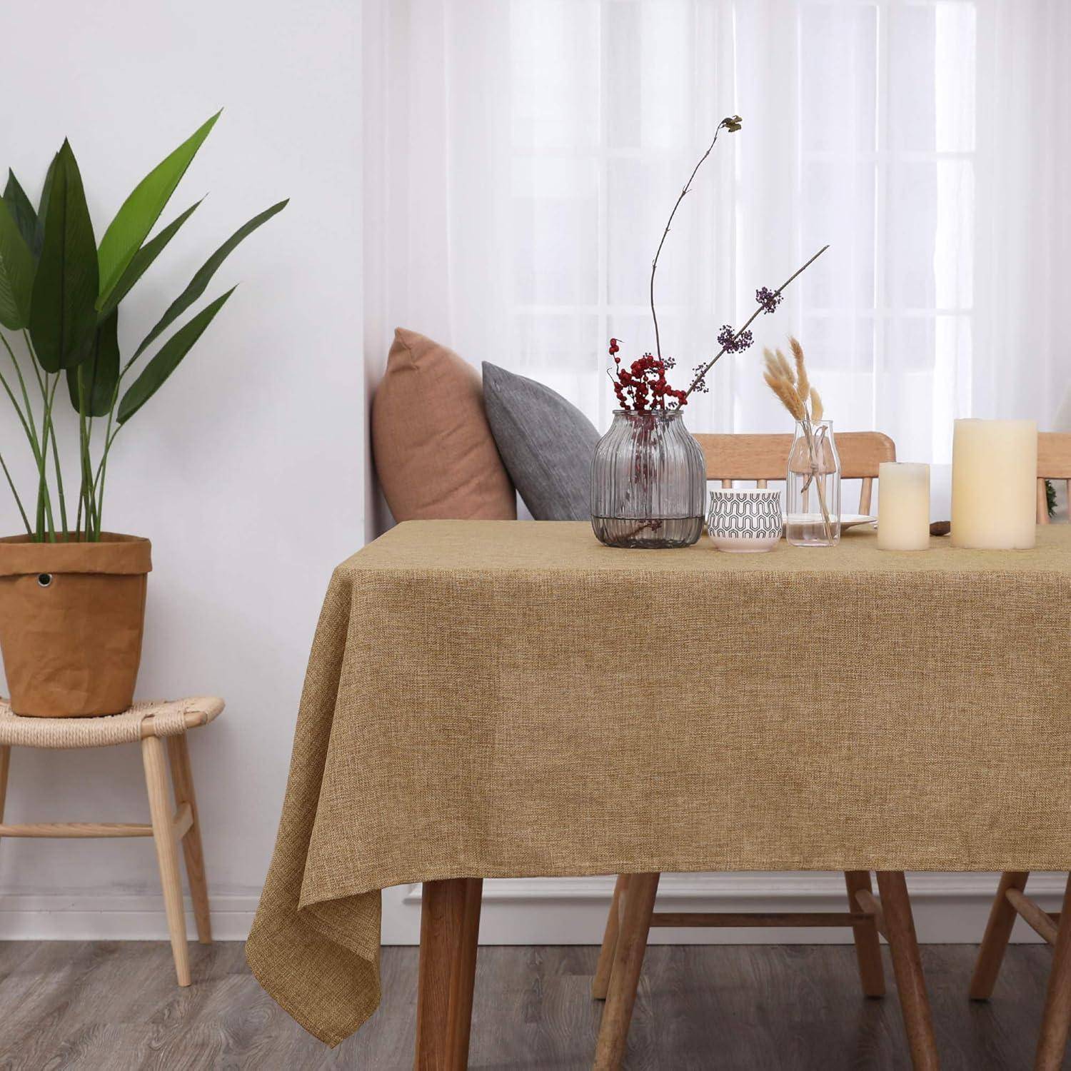 Ein minimalistisches Esszimmer mit einem Tisch, der mit einem beigen Tuch bedeckt ist, geschmückt mit Trockenzweigen in Vasen und brennenden Kerzen. Eine Pflanze und ein Stuhl befinden sich in der Nähe.