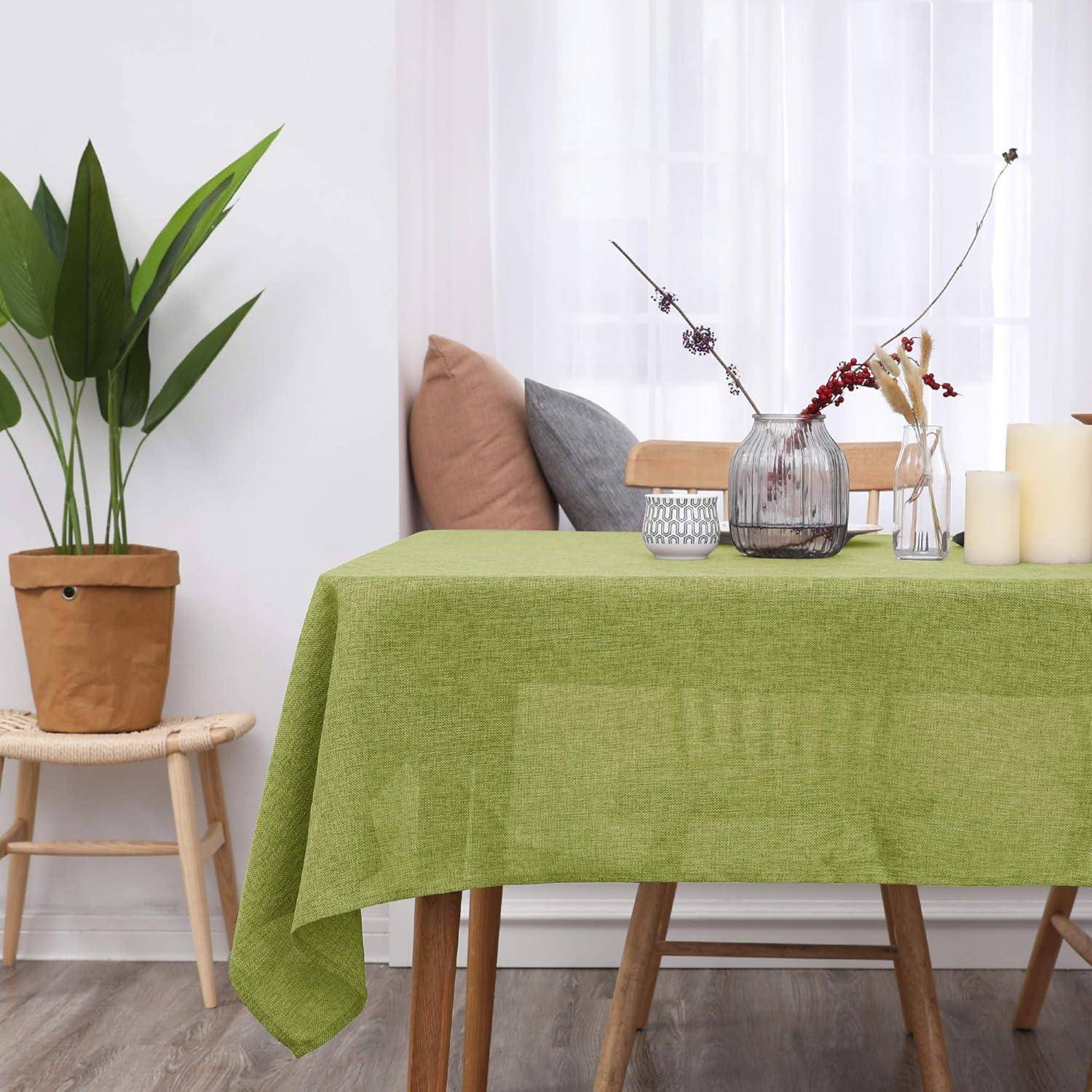 Ein gemütlicher Essbereich mit einem Holztisch, der mit einer grünen Tischdecke bedeckt ist, einer Vase mit Zweigen, Kerzen und einer Topfpflanze in der Nähe. Sanftes Licht fällt durch transparente Vorhänge.