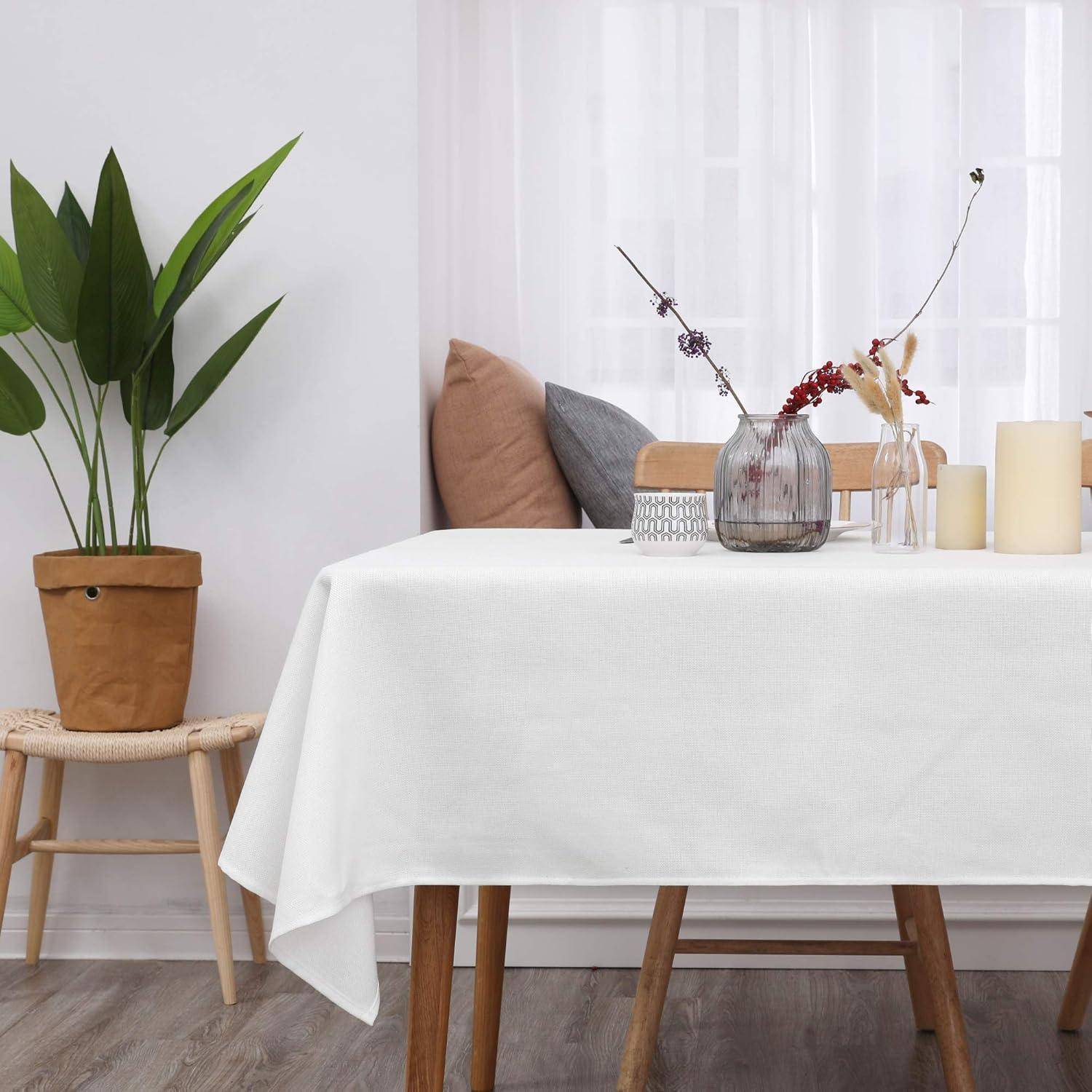 Ein Esstisch, bedeckt mit einer weißen Tischdecke, geschmückt mit einer Blumenvase, Kerzen und einer kleinen Pflanze auf einem geflochtenen Hocker in der Nähe.