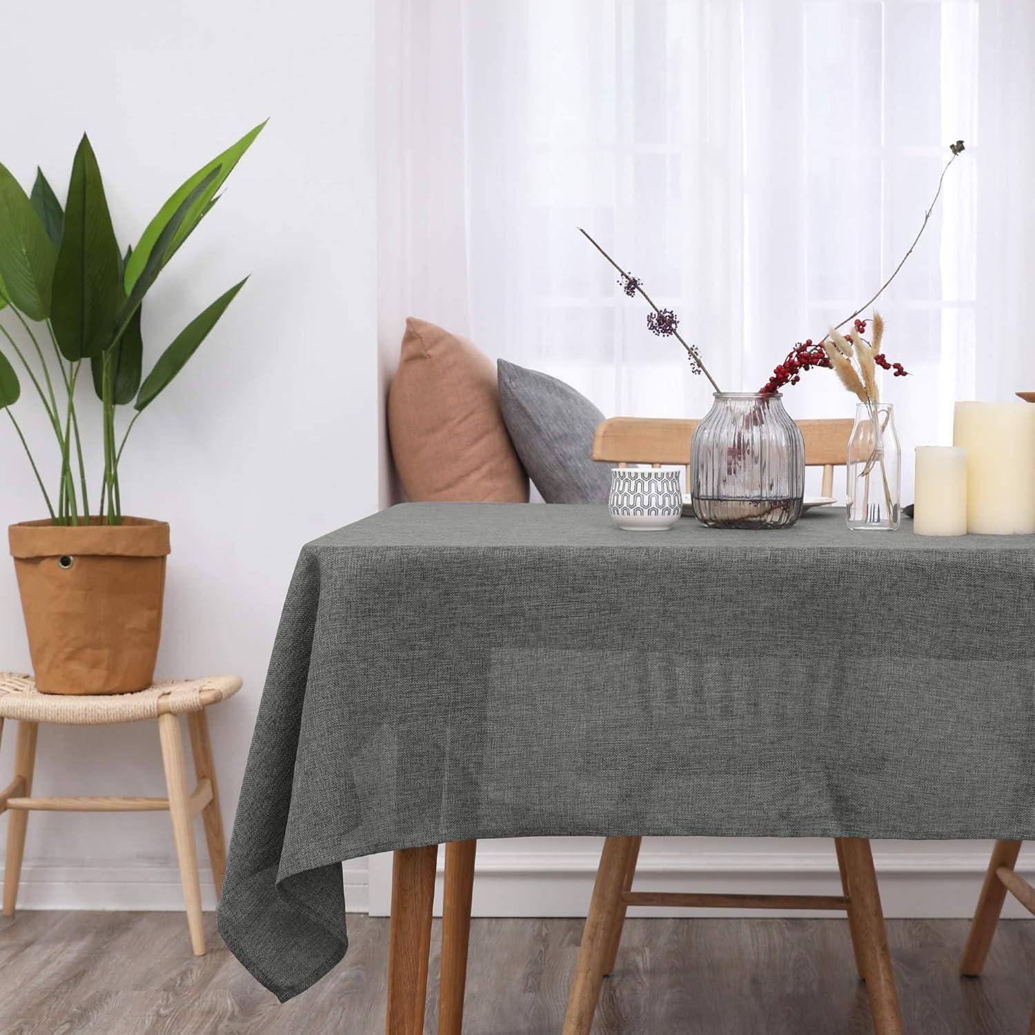 Ein Esstisch, der mit einer grauen Tischdecke bedeckt ist, trägt eine Vase mit lila Blumen, eine Kerze und einen kleinen Blumentopf. Daneben steht ein Pflanze auf einem Hocker.
