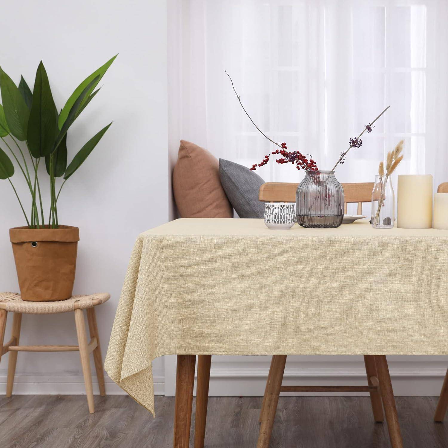 Ein Tisch mit einer beigen Tischdecke trägt dekorative Gegenstände wie eine Vase mit Zweigen, Kerzen und eine Pflanze. Ein Stuhl und eine Topfpflanze befinden sich in der Nähe.