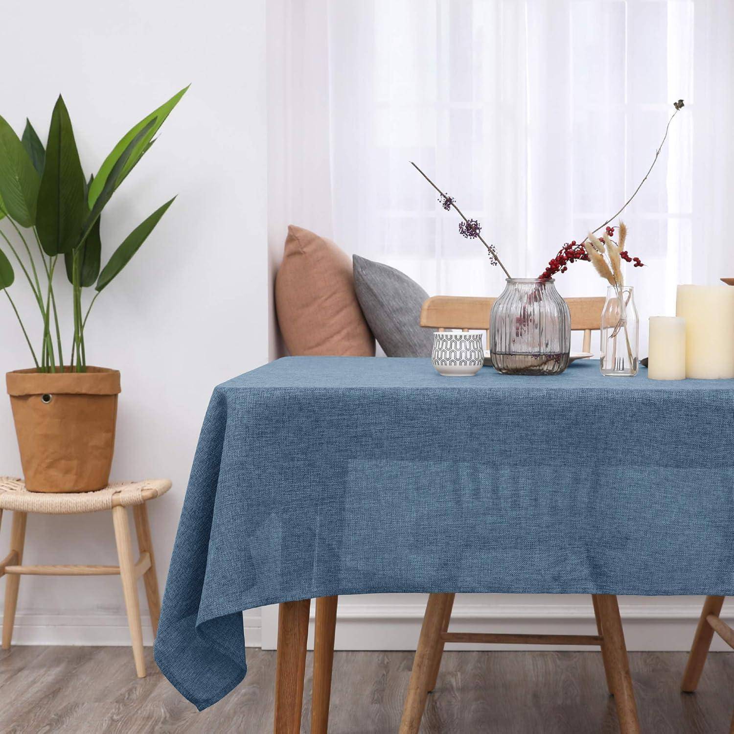 Ein Tisch mit einer blauen Decke ist mit Kerzen, Vasen und einer Pflanze auf einem nahegelegenen Hocker gedeckt und schafft eine schlichte, elegante Dekorszene.