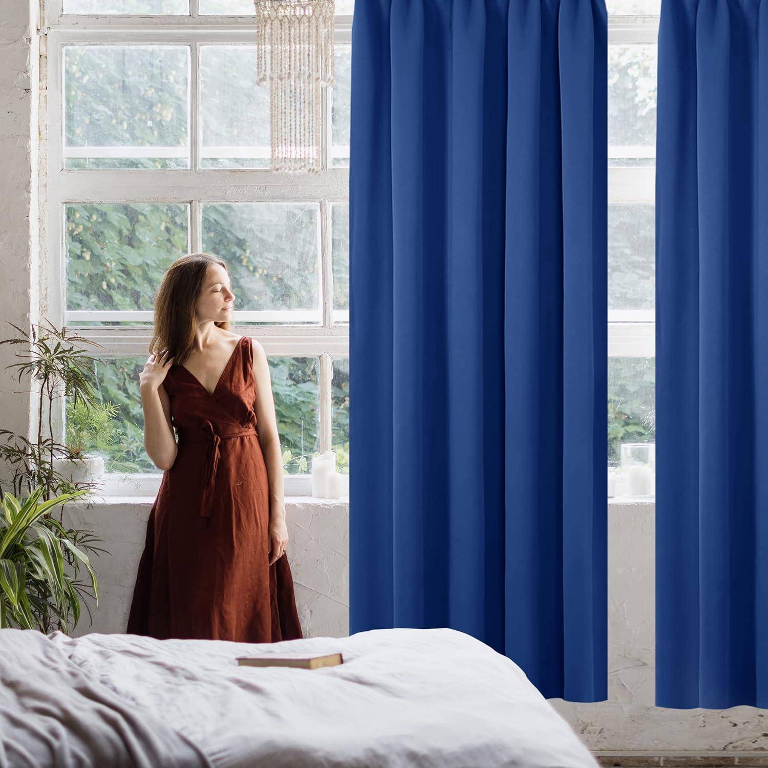 Eine Frau in einem langen braunen Kleid steht an einem großen Fenster mit blauen Vorhängen und blickt nach draußen. Zimmerpflanzen und ein Kronleuchter vervollständigen die Dekoration.