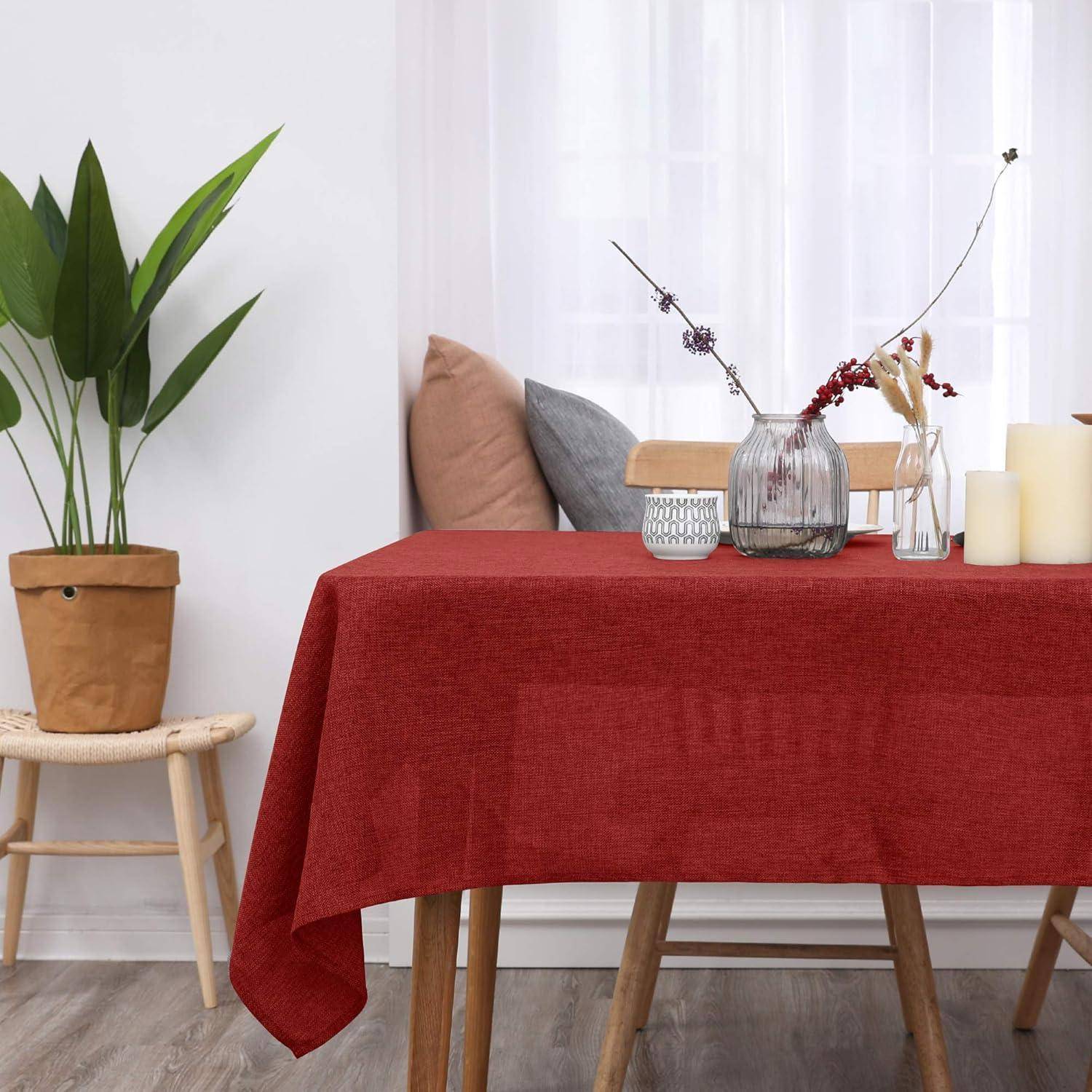 Eine rote Tischdecke bedeckt einen Esstisch mit einer dekorativen Vase und Kerzenarrangement; eine Topfpflanze und ein gepolsterter Stuhl befinden sich in der Nähe.