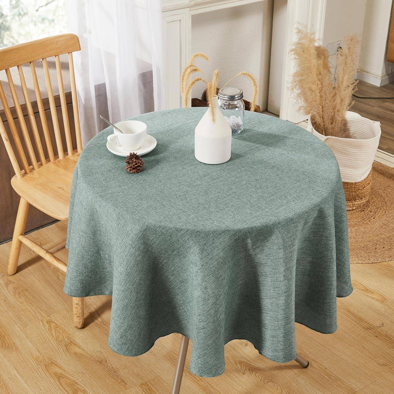 Ein runder Tisch mit einer türkisfarbenen Tischdecke, darauf eine weiße Tasse mit Untertasse, ein silberner Milchkrug und ein Tannenzapfen. Ein Holzstuhl steht in der Nähe.