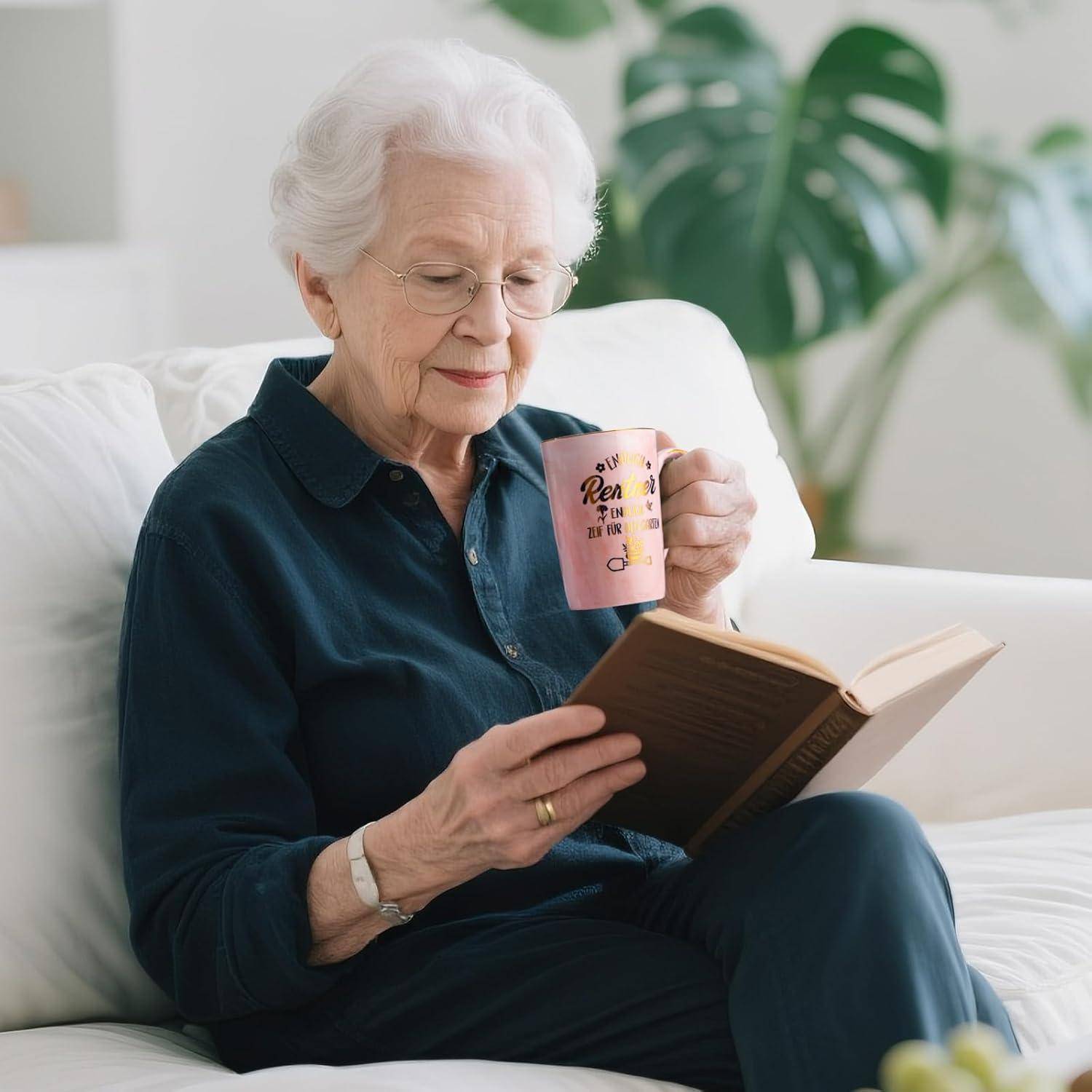 Eine ältere Frau sitzt auf einem weißen Sofa, liest ein Buch und hält eine rosa Tasse. Sie wirkt entspannt und konzentriert in einem hellen Raum.