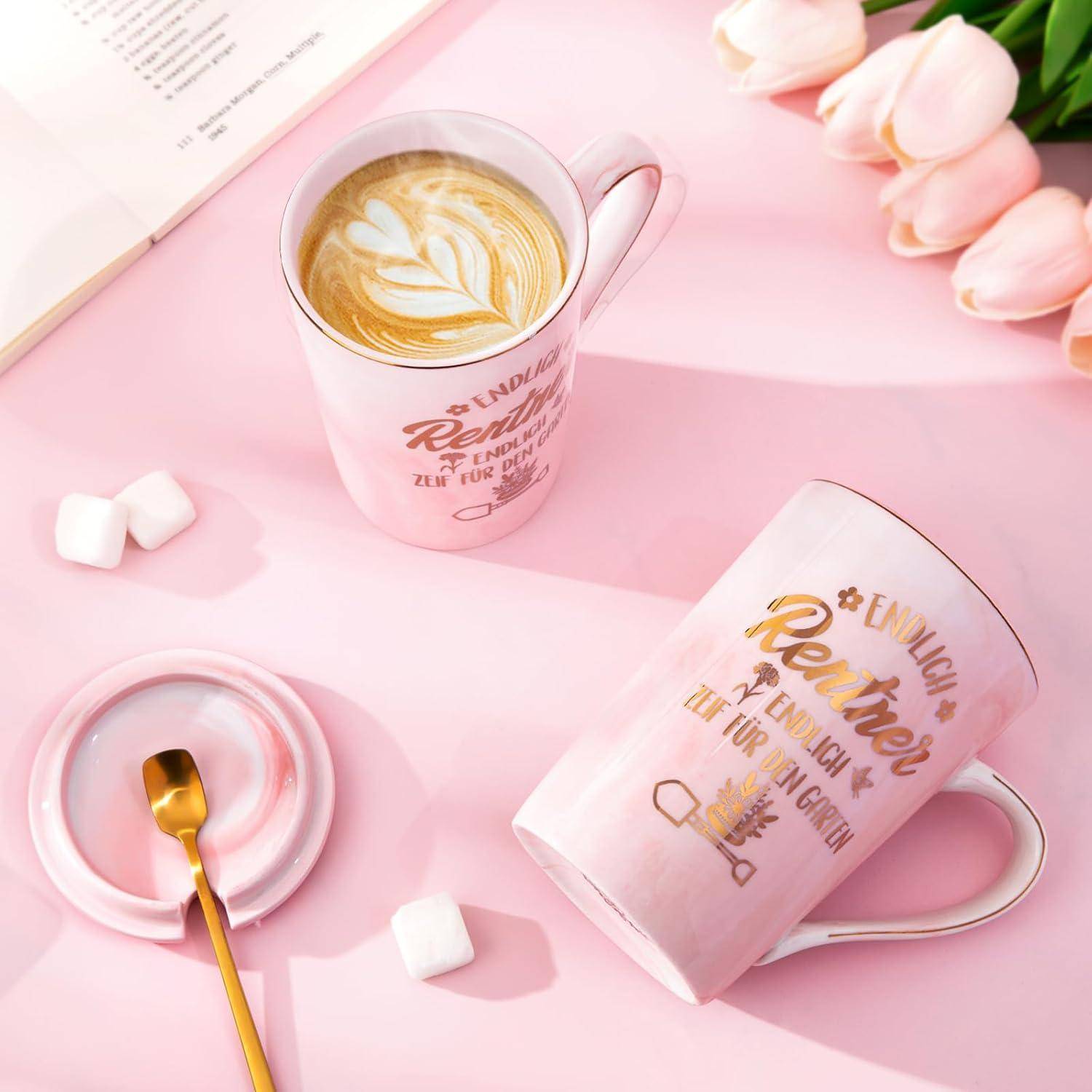 Eine rosa Tasse mit Latte-Kunst auf der Oberfläche steht auf einer passenden Untertasse. Ein aufgeschlagenes Buch und Marshmallows befinden sich in der Nähe auf einer rosa Oberfläche.