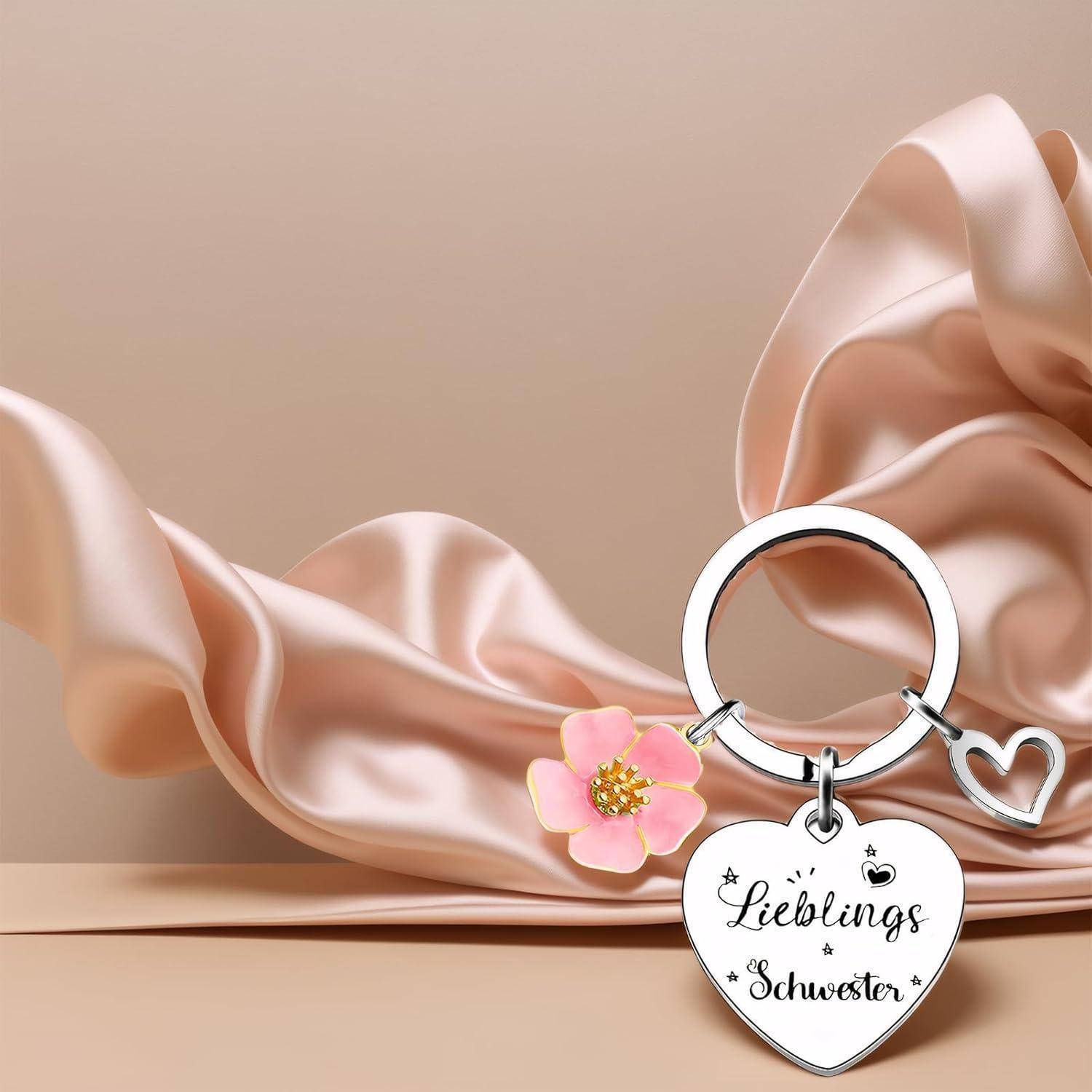 Eine herzförmige Schlüsselanhänger mit der Aufschrift 'Lieblings Schwester' und einem kleinen pinken Blumendesign liegt auf rosa Satin-Stoff.