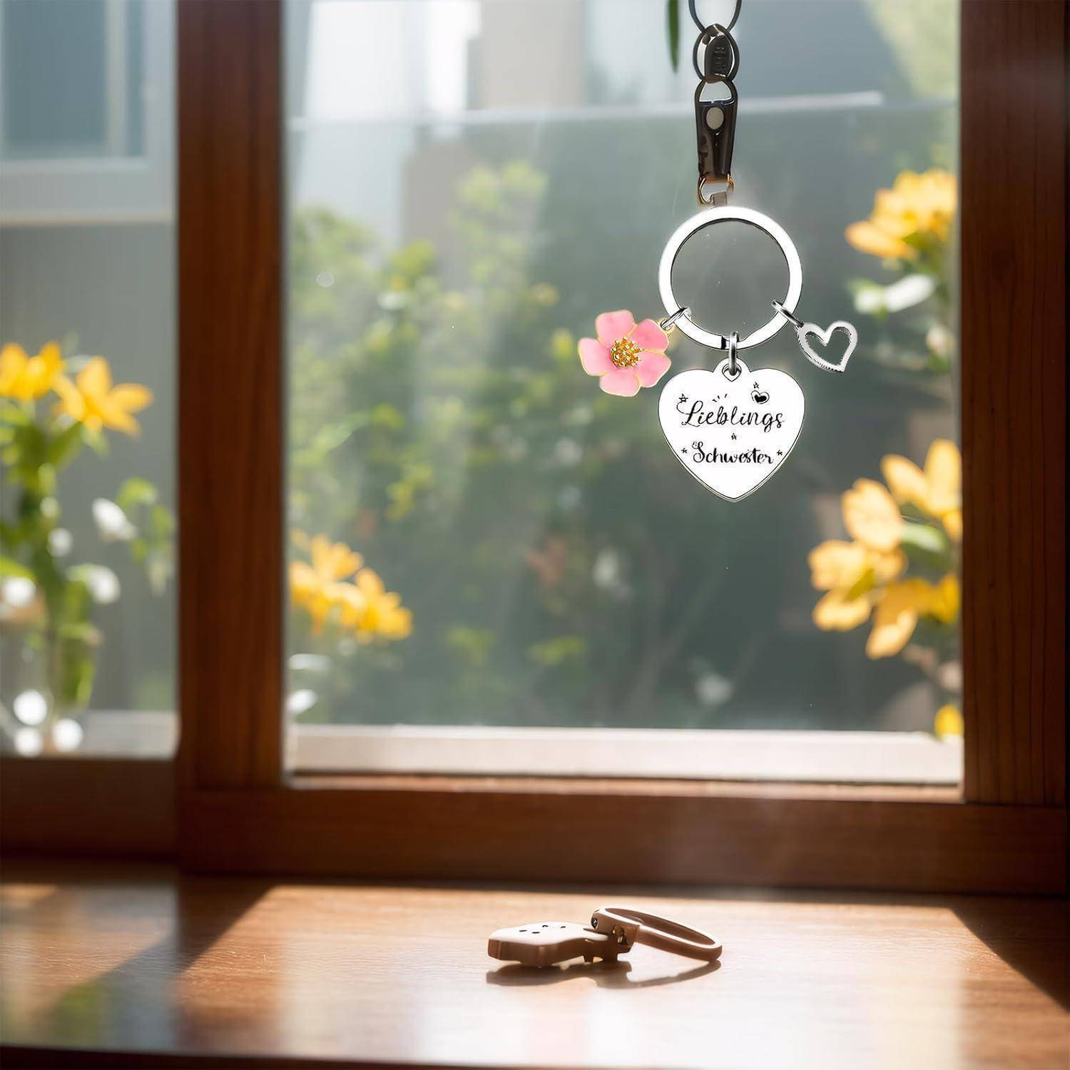 Ein herzförmiger Schlüsselanhänger mit der Aufschrift 'Lieblings Schwester' und einem pinken Blumenanhänger hängt vor einem Fenster, mit Sonnenlicht und Blumen im Hintergrund.