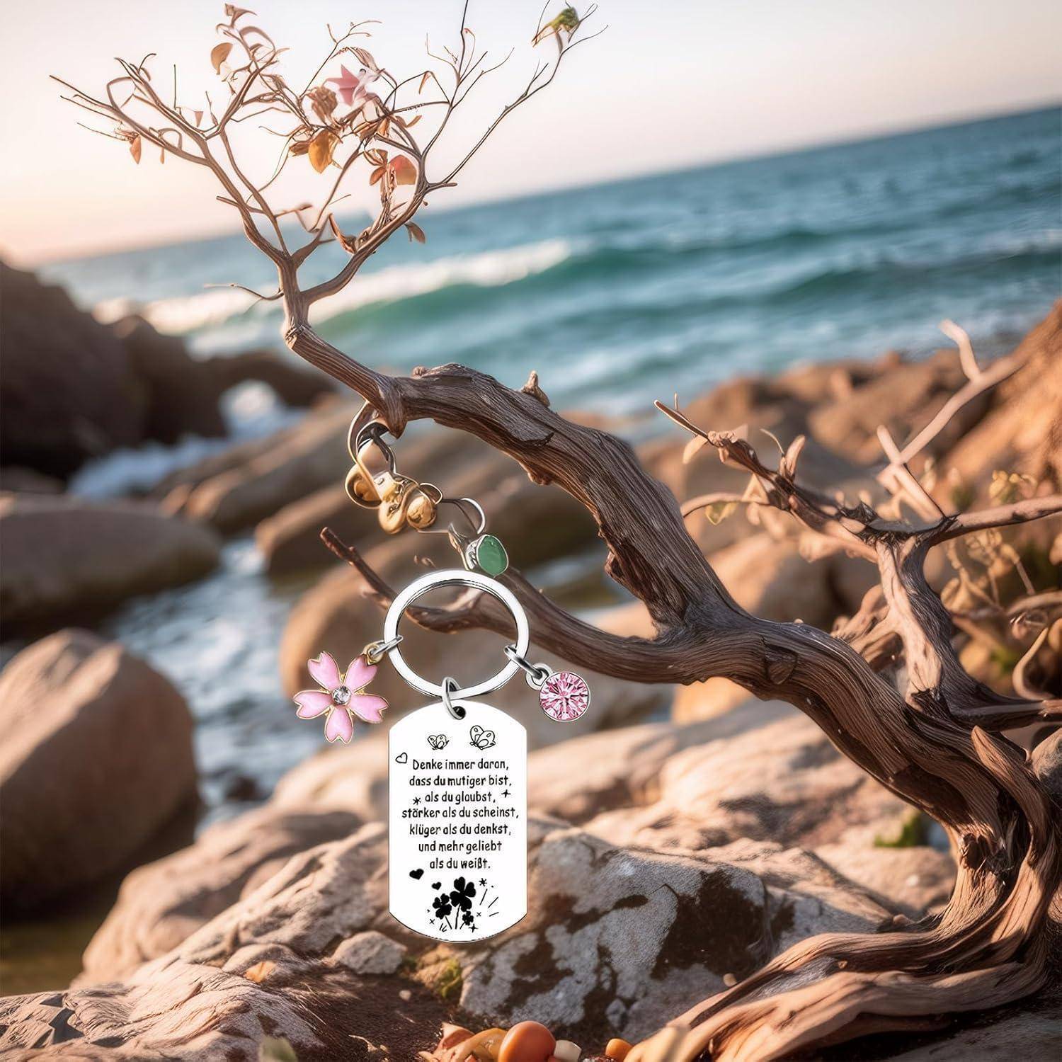 Ein Schlüsselanhänger mit Anhängern hängt an einem kleinen Baumast am Meer. Die Landschaft zeigt Felsen und Wasser unter einem klaren Himmel bei Sonnenuntergang.